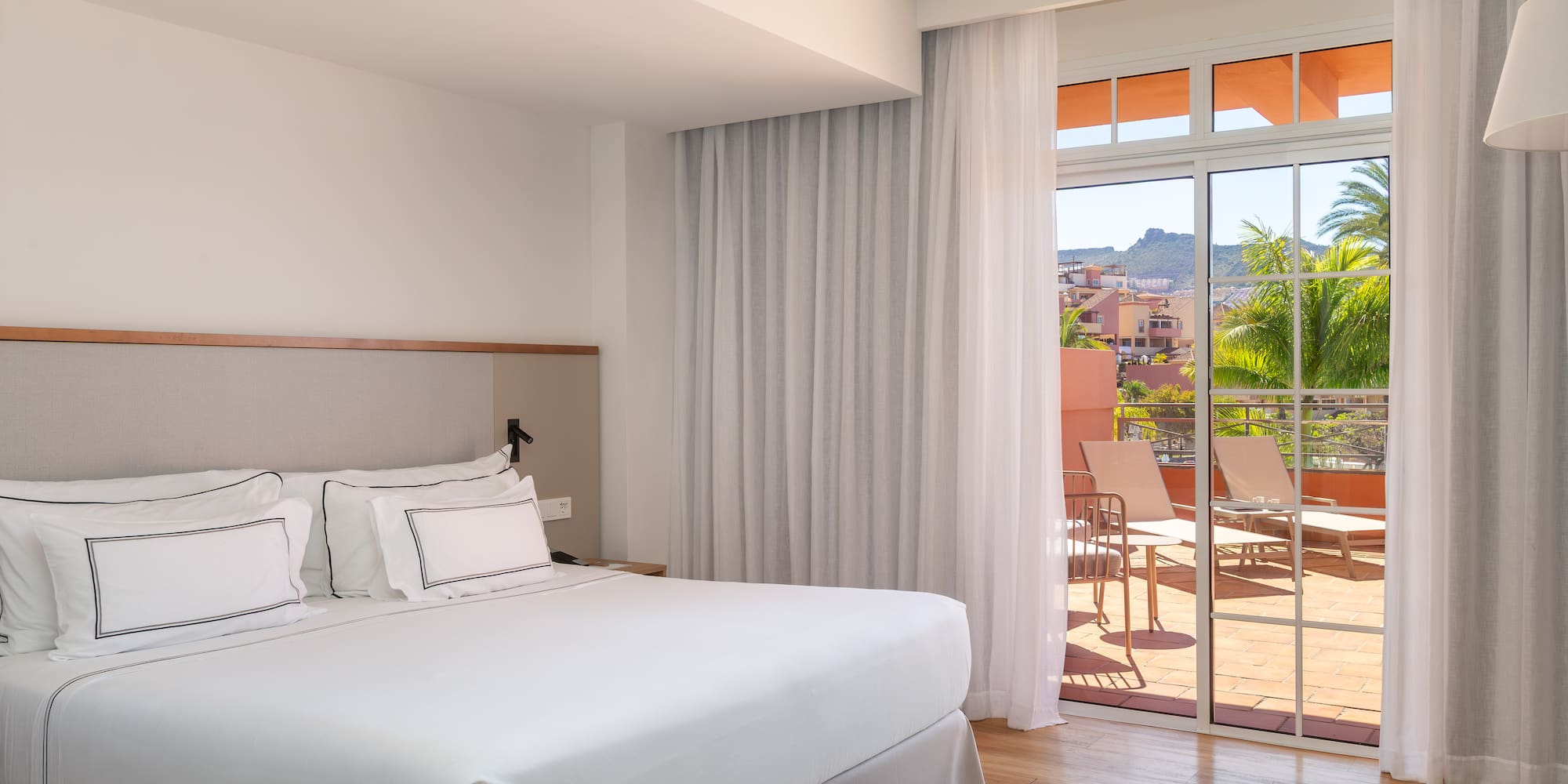 Bright hotel bedroom with king bed and balcony overlooking tropical resort view.