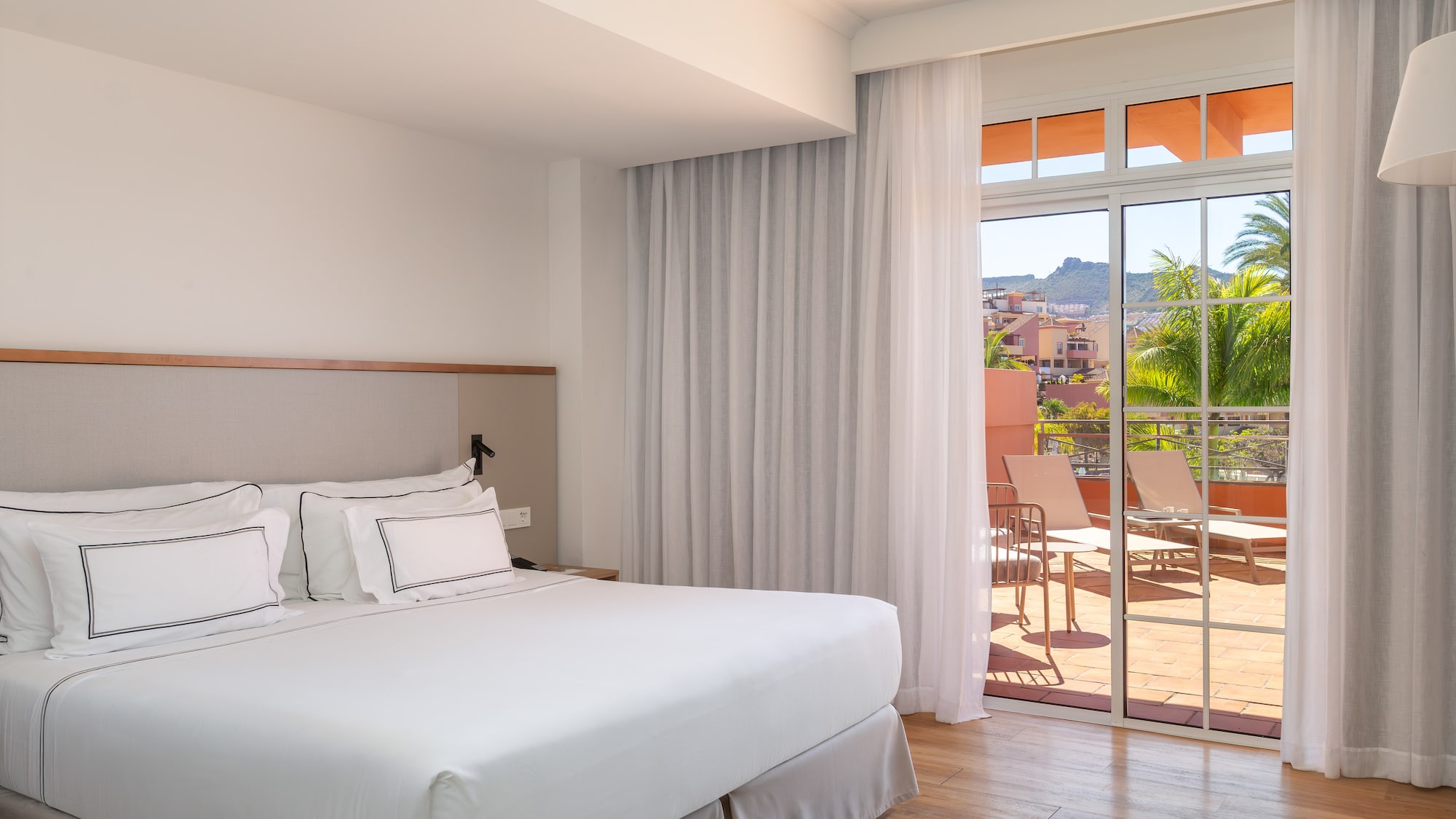 Bright hotel bedroom with king bed and balcony overlooking tropical resort view.