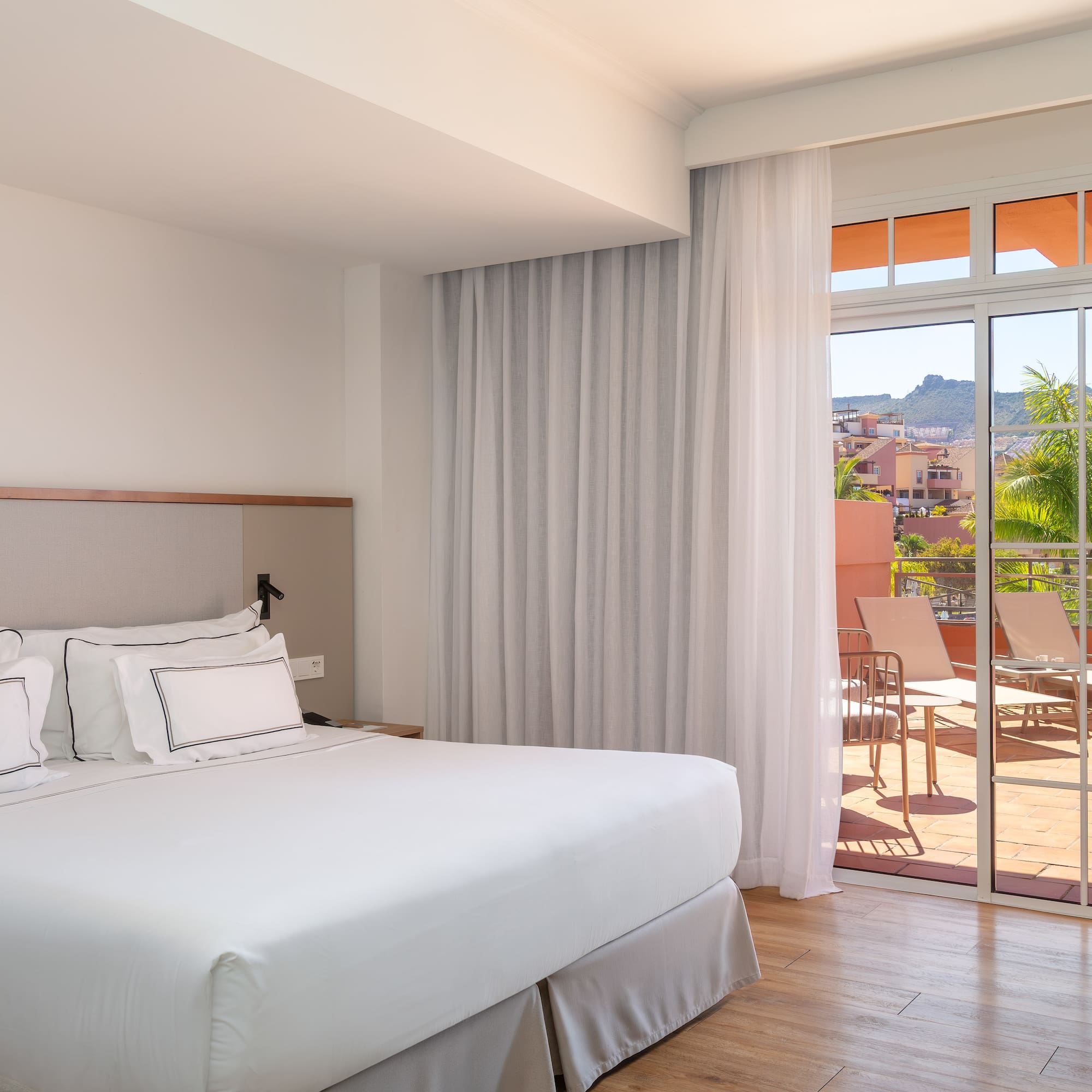 Bright hotel bedroom with king bed and balcony overlooking tropical resort view.