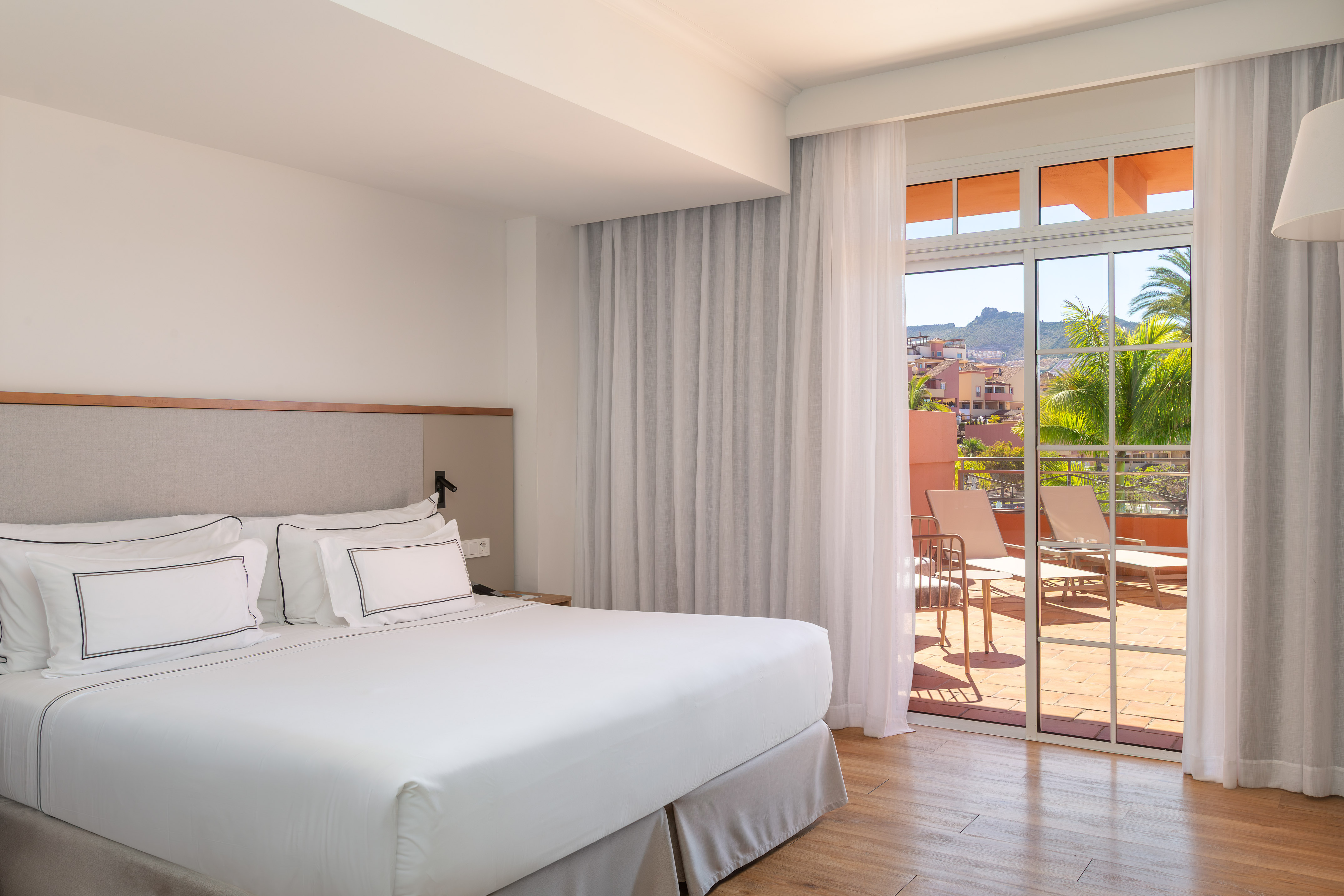 Bright hotel bedroom with king bed and balcony overlooking tropical resort view.