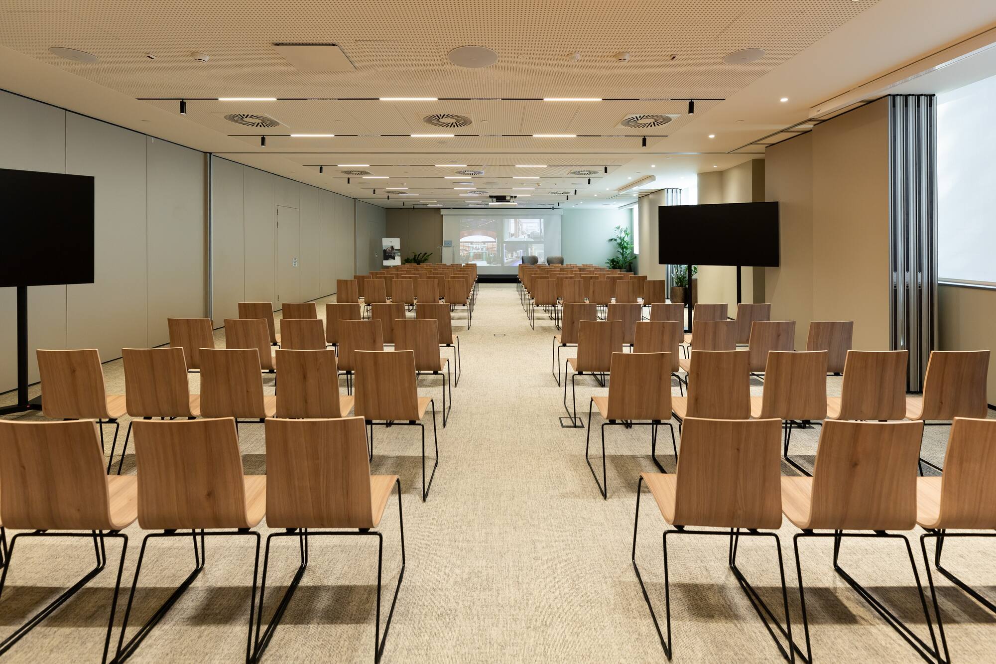 Modern conference room with rows of wooden chairs, large screen, and monitors.