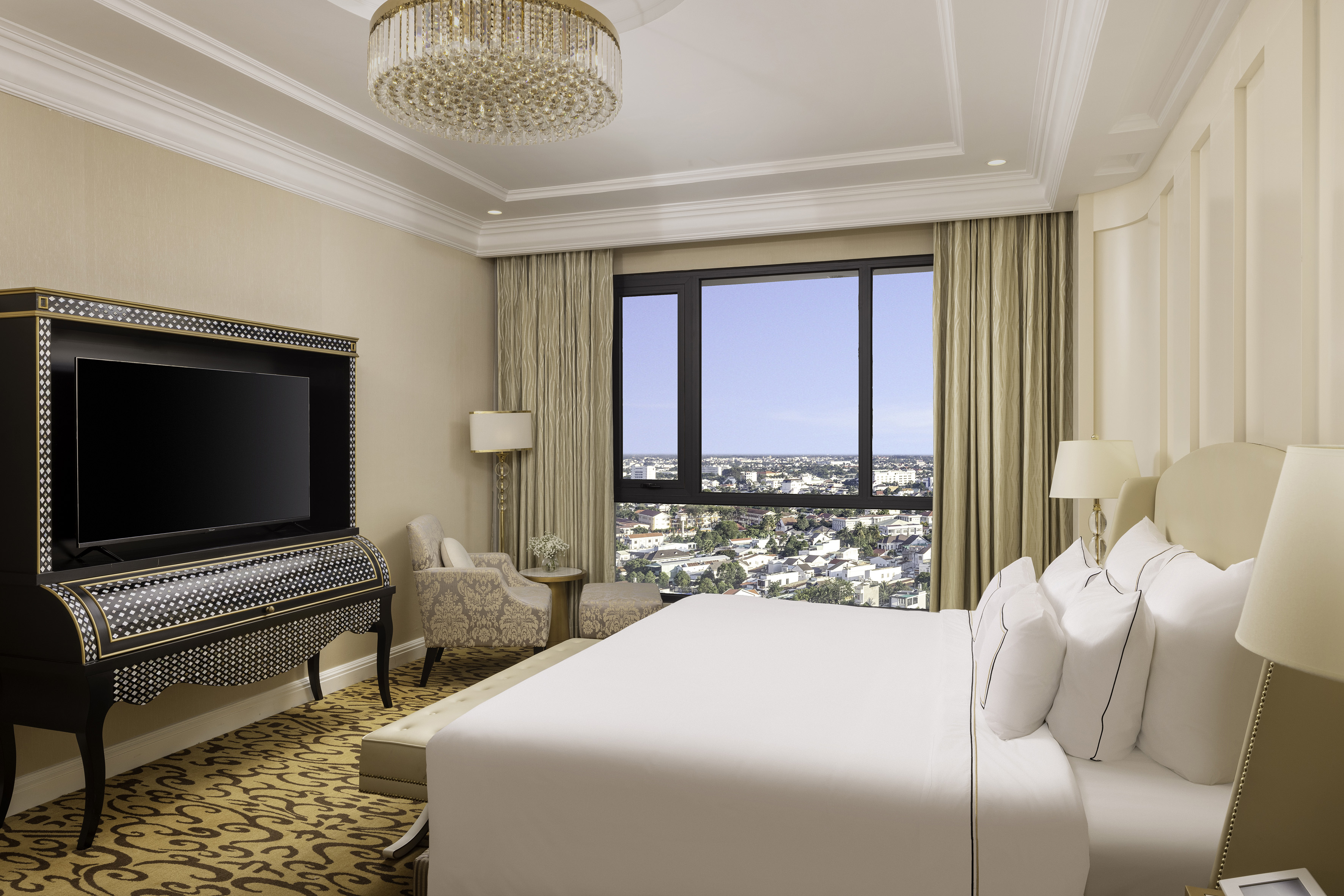 Luxury hotel room with city view, large bed, ornate cabinet, and chandelier.