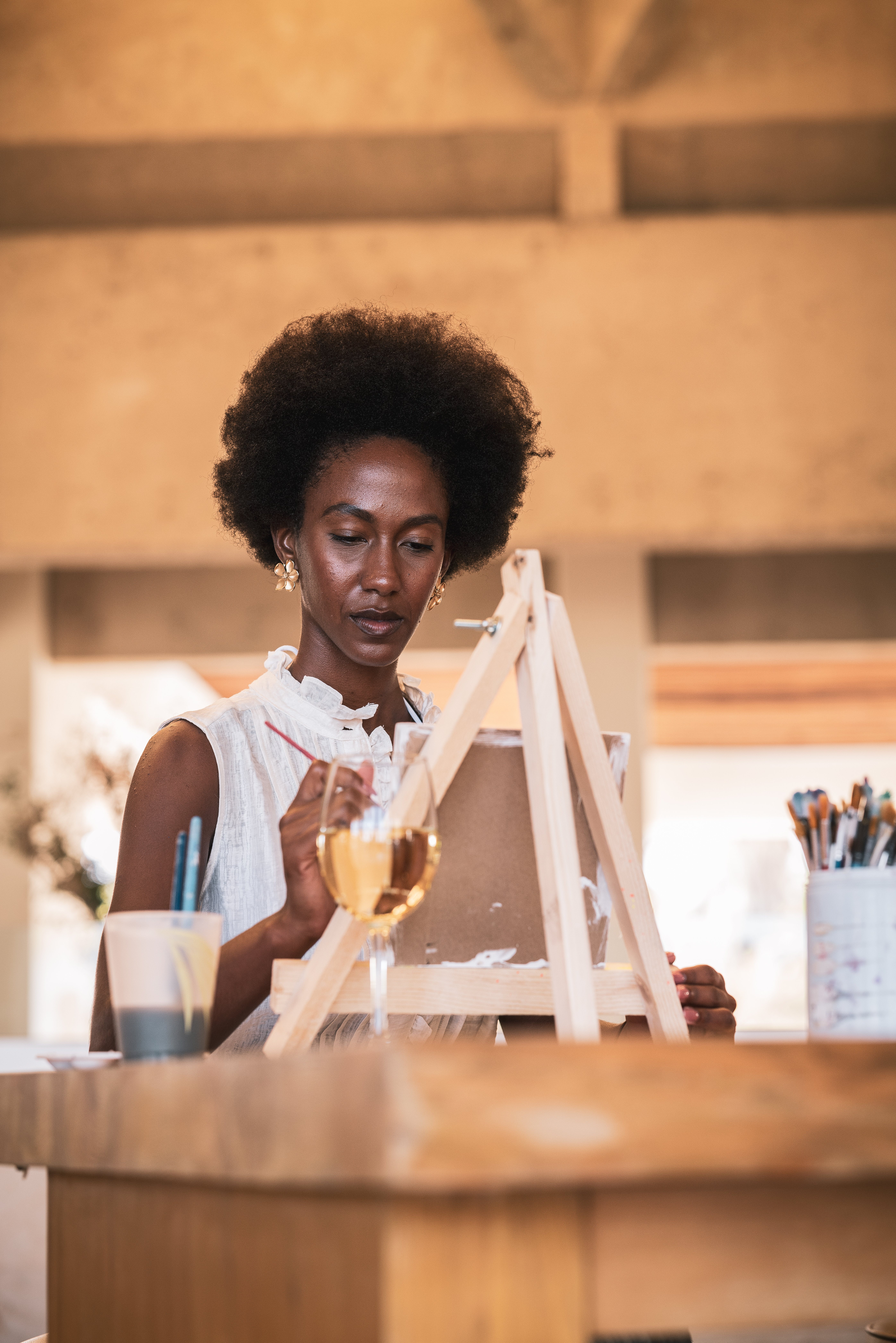 Afro woman artist painting on easel with brush and wine glass.