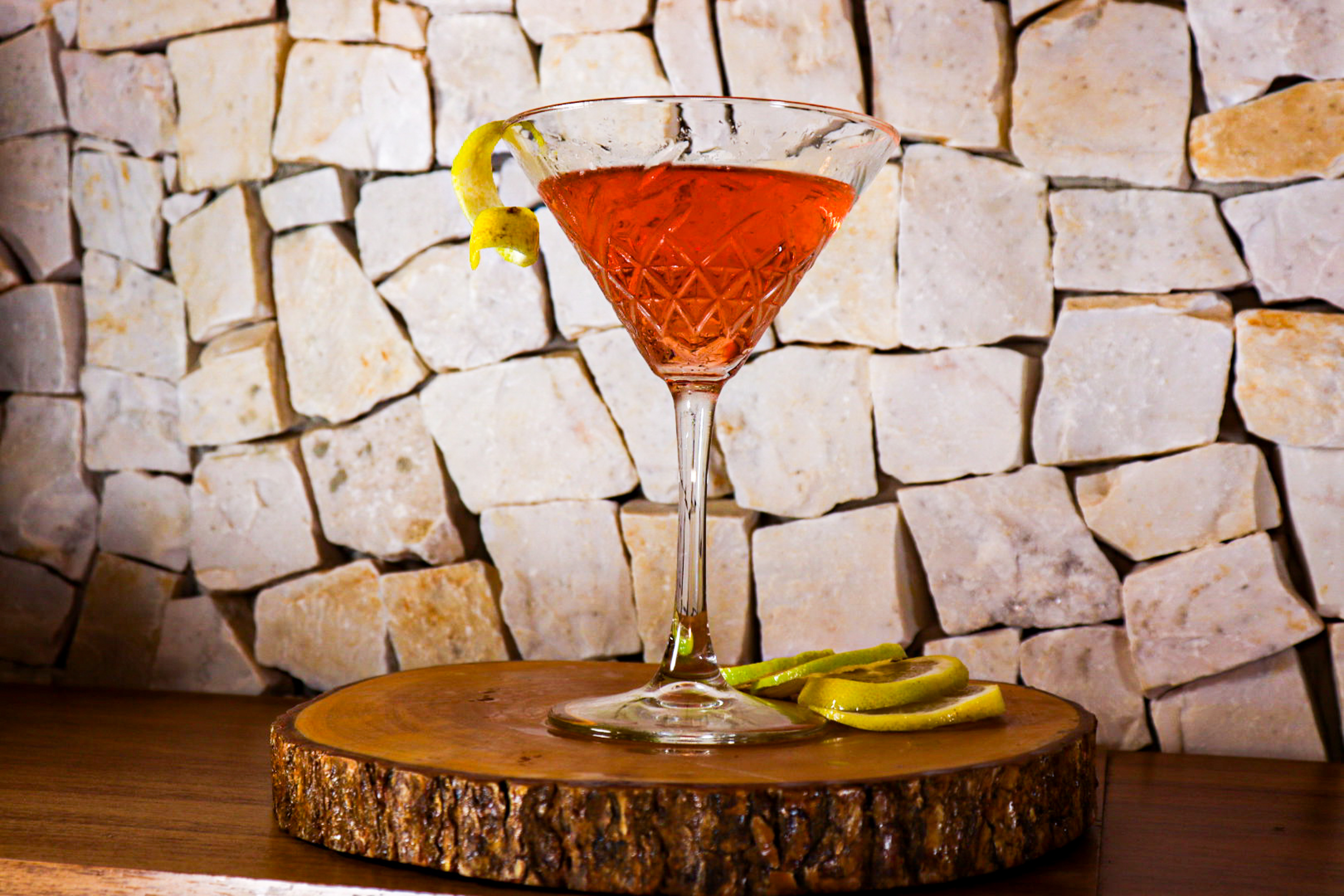 Red cocktail in martini glass with lemon peel, on wood slice, stone wall.