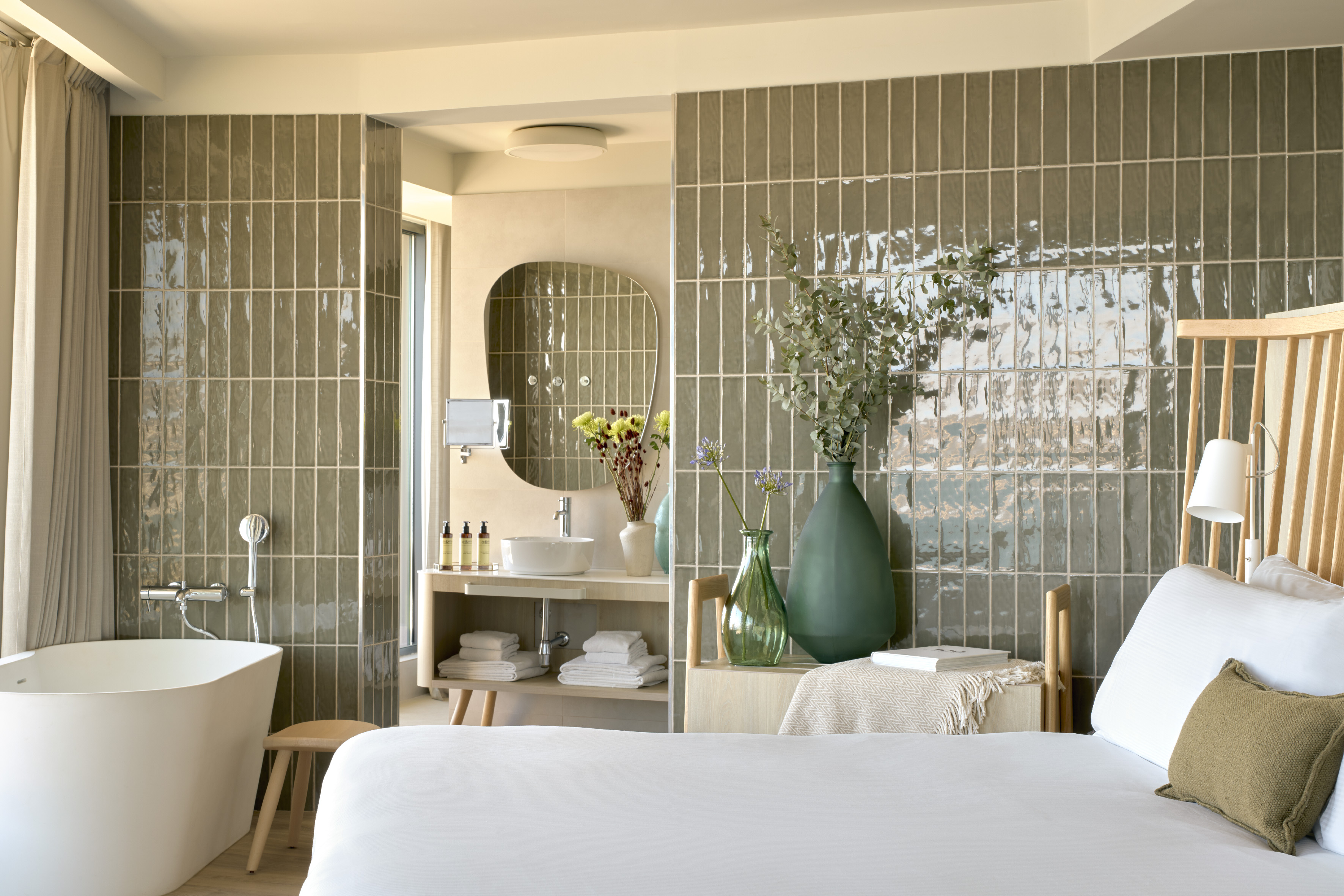 Modern hotel suite with freestanding tub, green subway tiles, and white bed.
