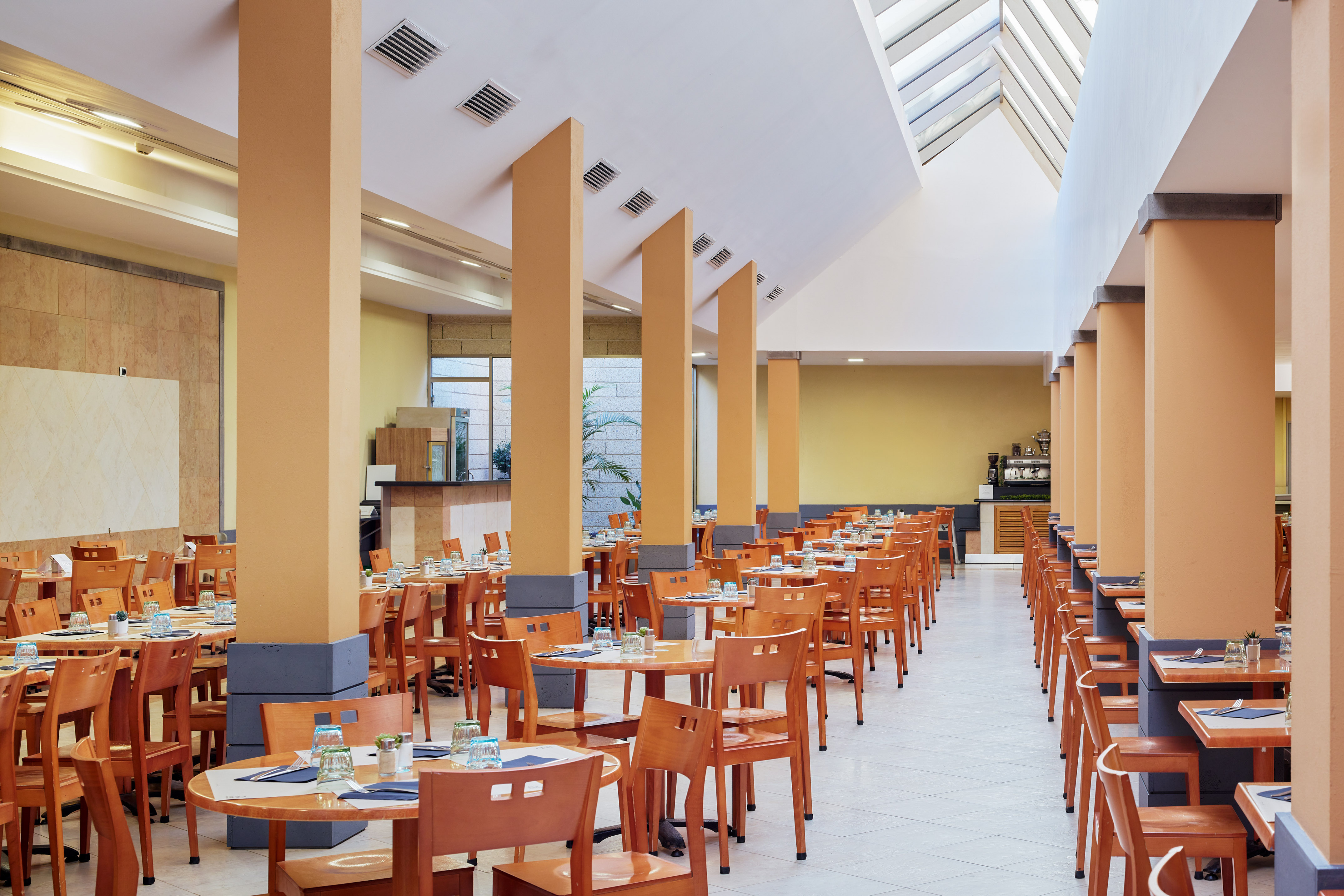 Bright hotel dining room with many wooden tables, chairs, and tall columns.