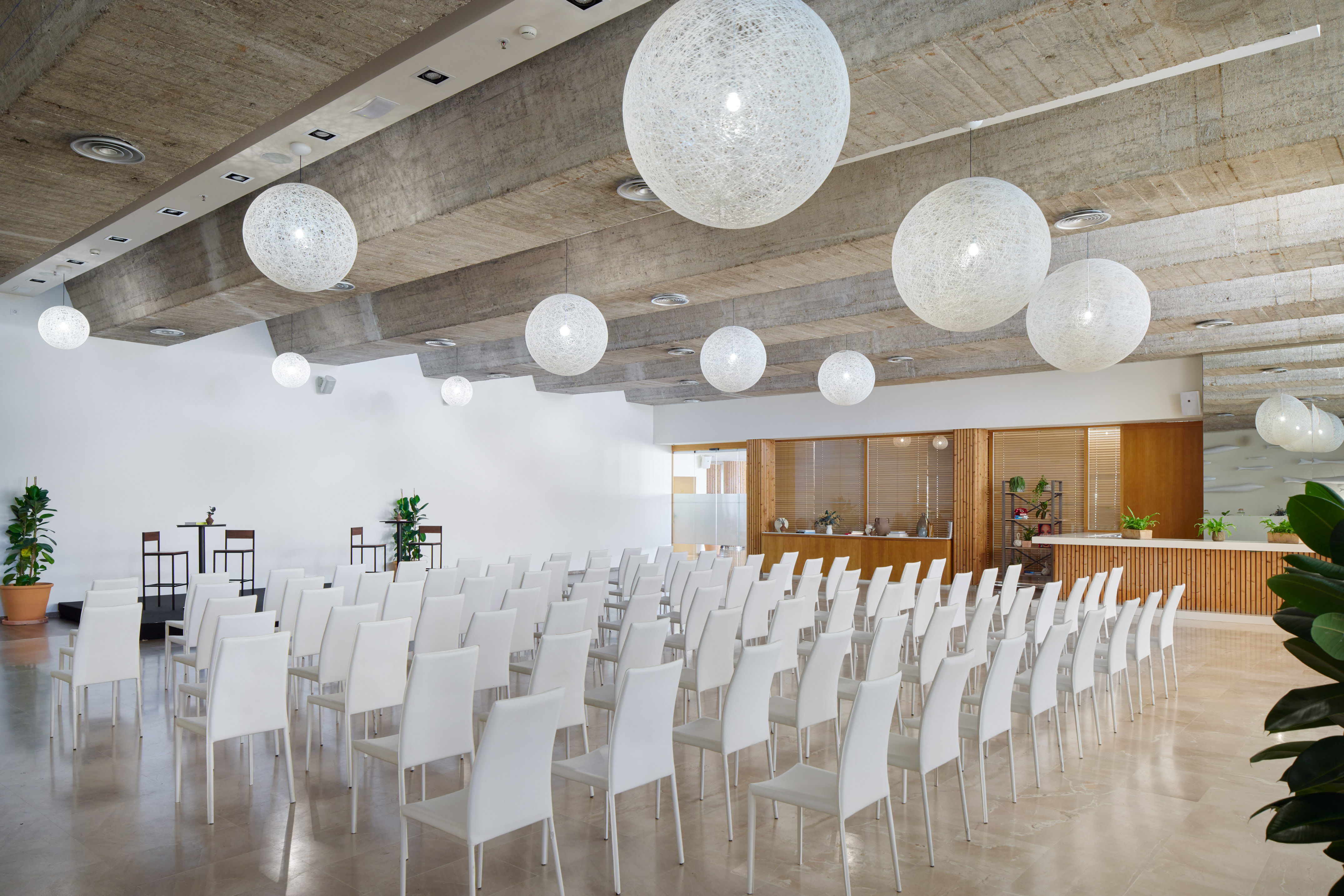 Modern event hall with white chairs, spherical pendant lights, and concrete ceiling.