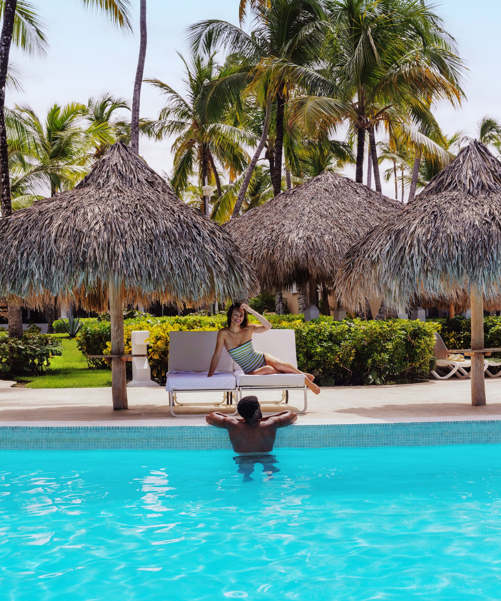 Couple by tropical resort pool; woman on lounge, man in water, palm trees.
