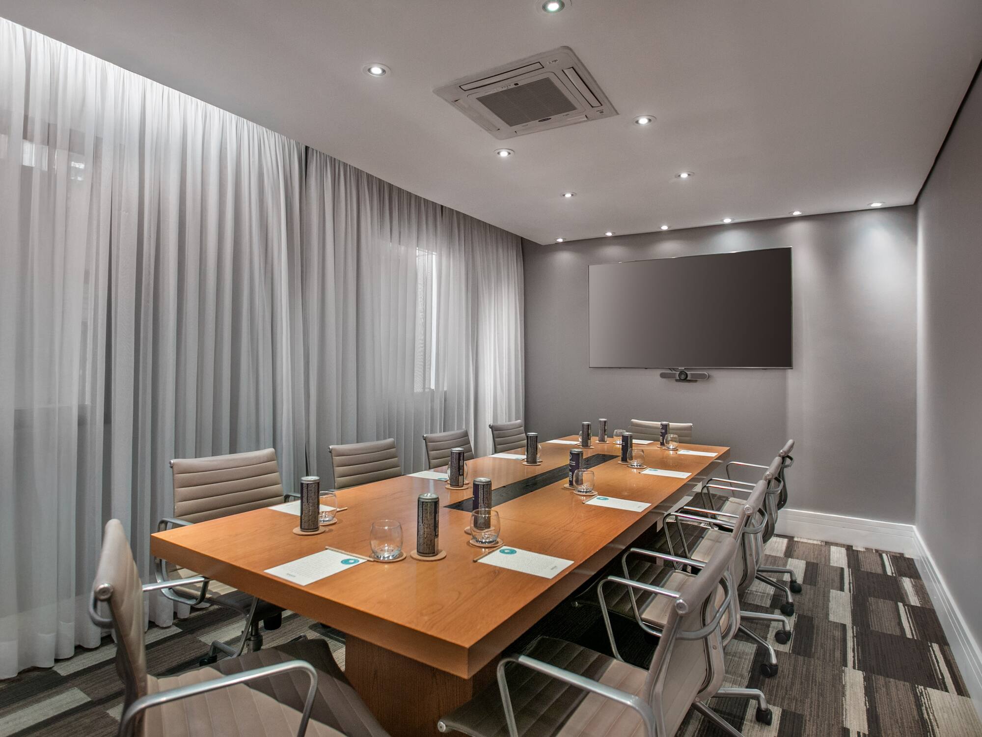 Modern conference room with long wood table, gray chairs, large screen, and curtains.