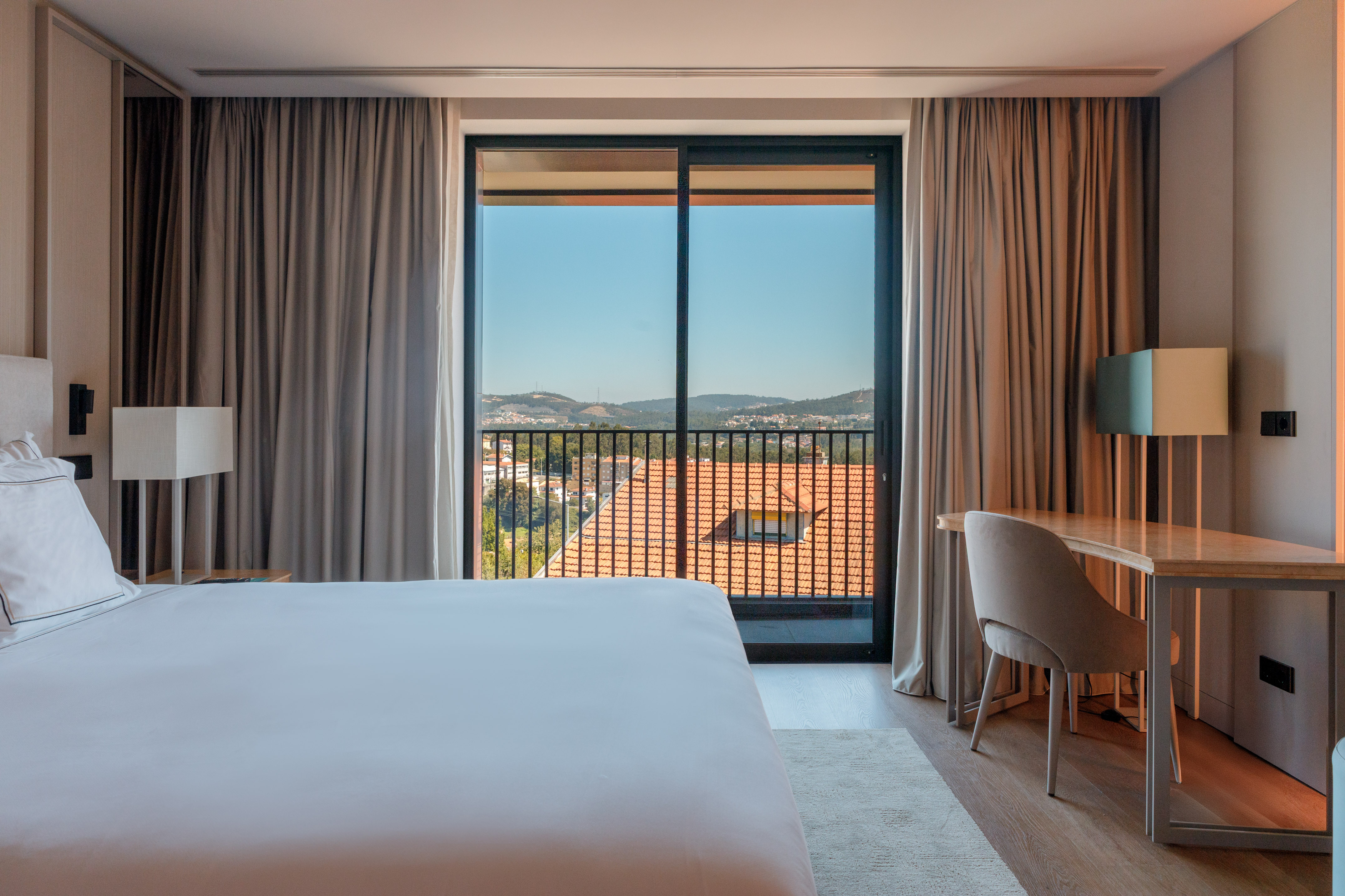 Modern hotel bedroom with balcony city view, white bed, and desk area.