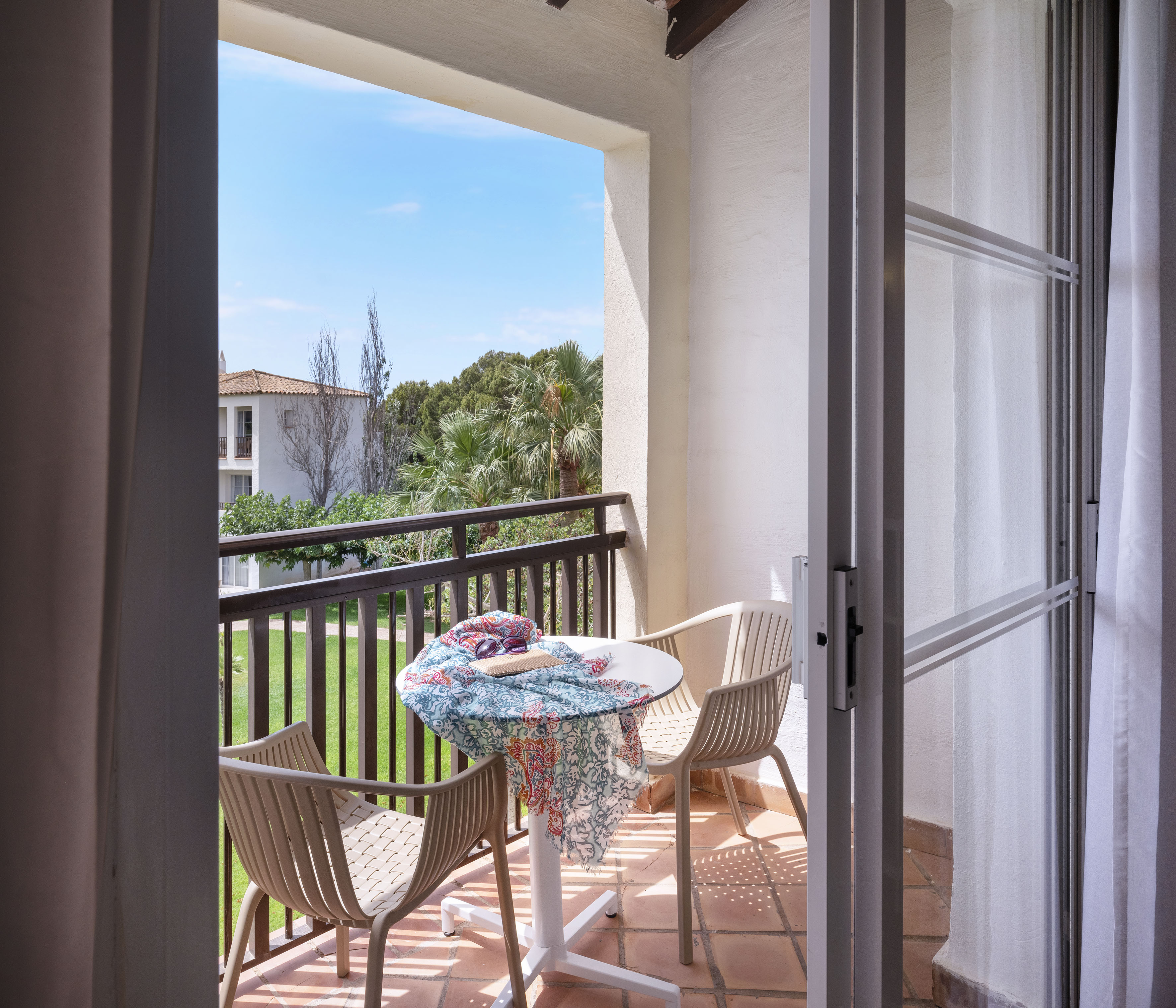 Sunny hotel balcony with outdoor seating, garden view, and palm trees.