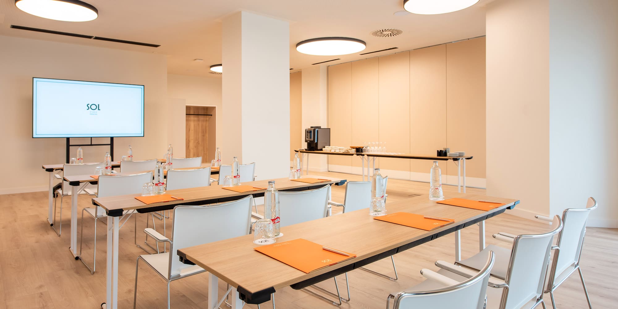 Modern meeting room with presentation screen, tables, white chairs, and coffee station.