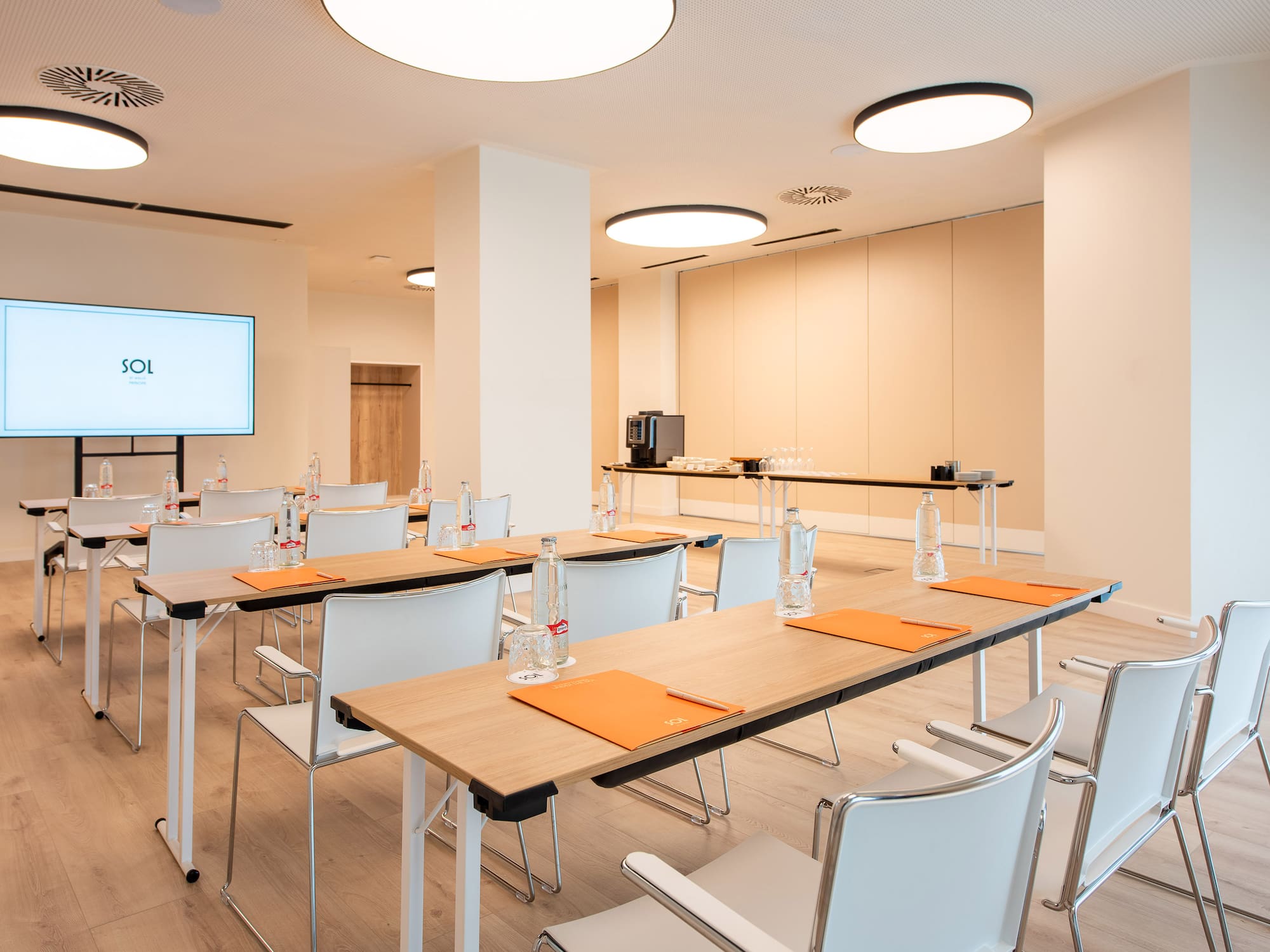 Bright, modern meeting room with seminar tables, white chairs, large screen, and refreshments.