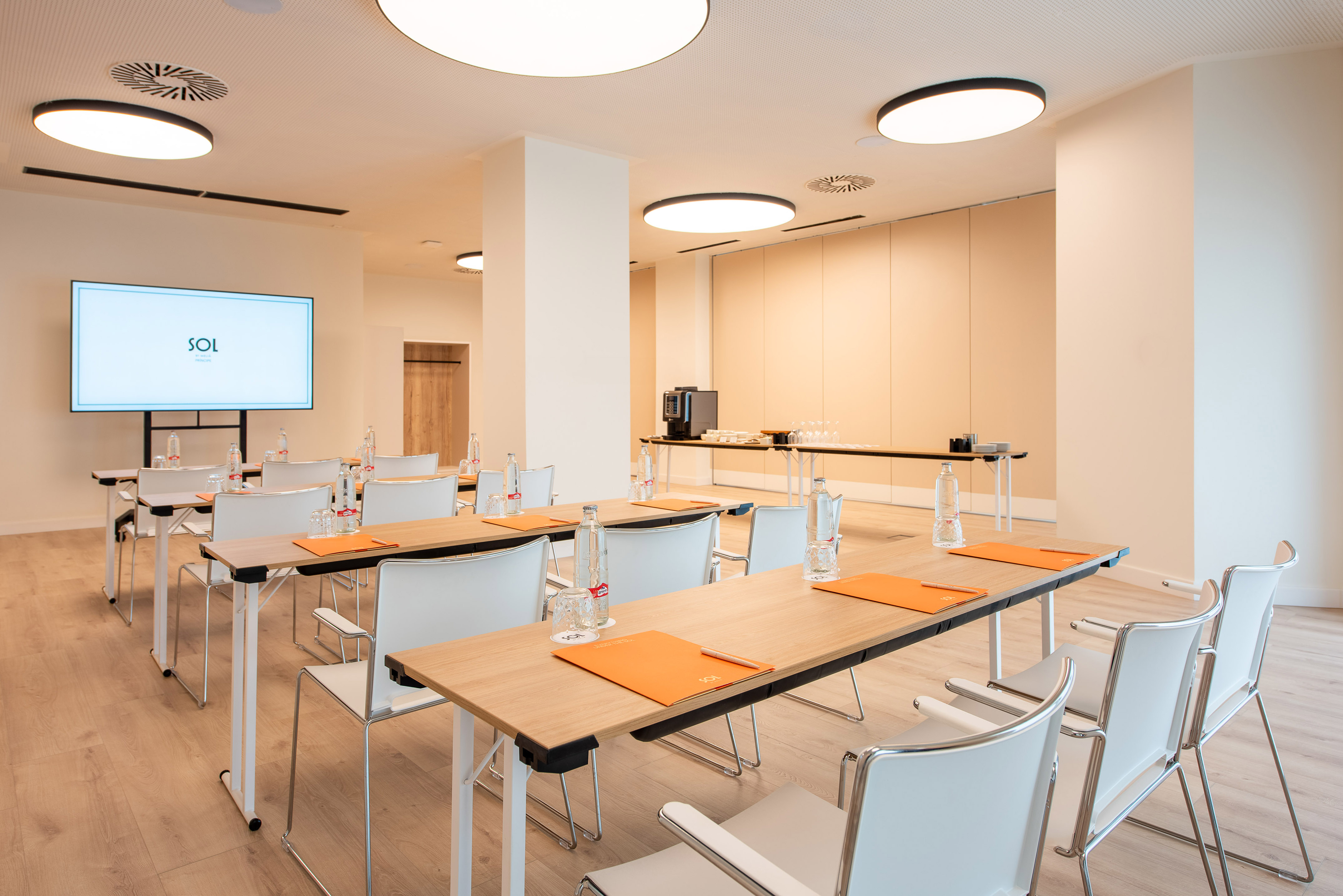 Bright, modern meeting room with seminar tables, white chairs, large screen, and refreshments.