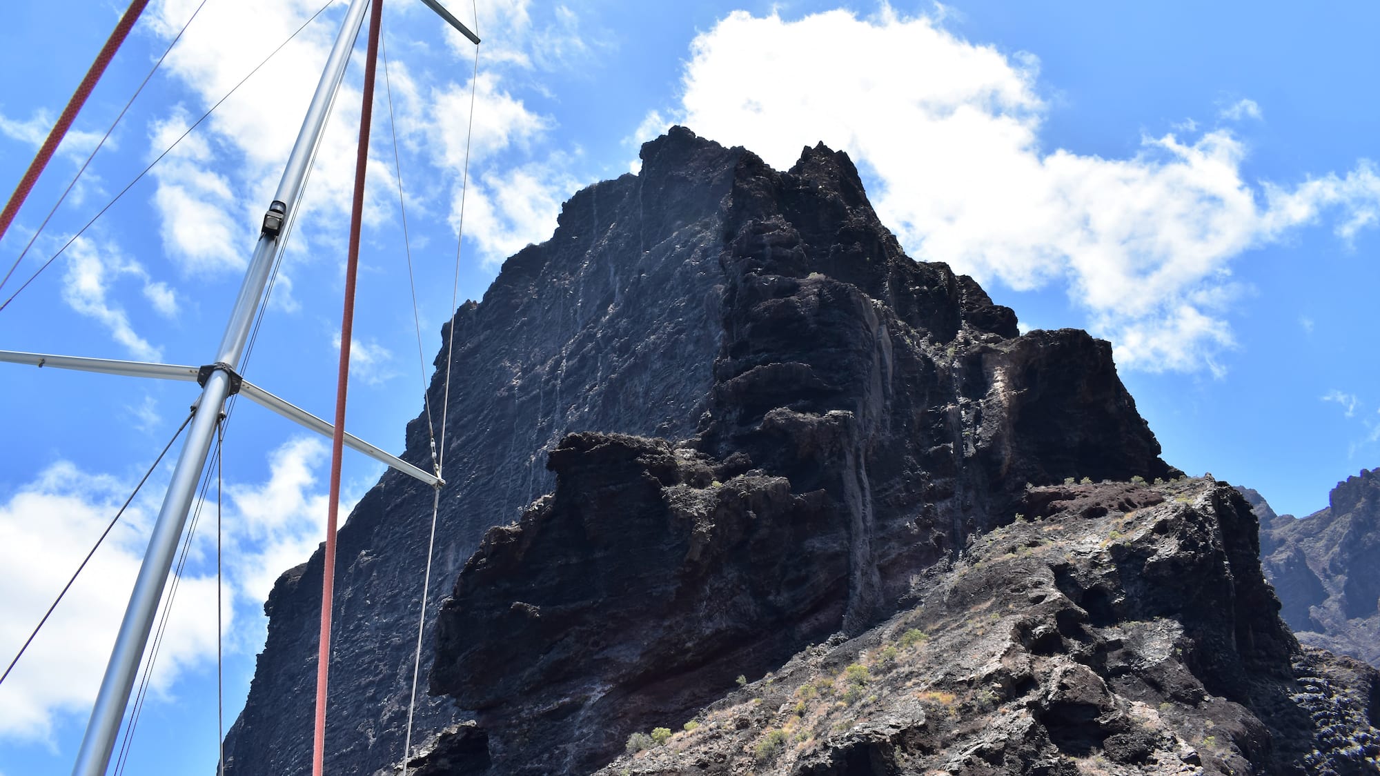 Dark volcanic cliff and sailboat mast under cloudy blue sky.