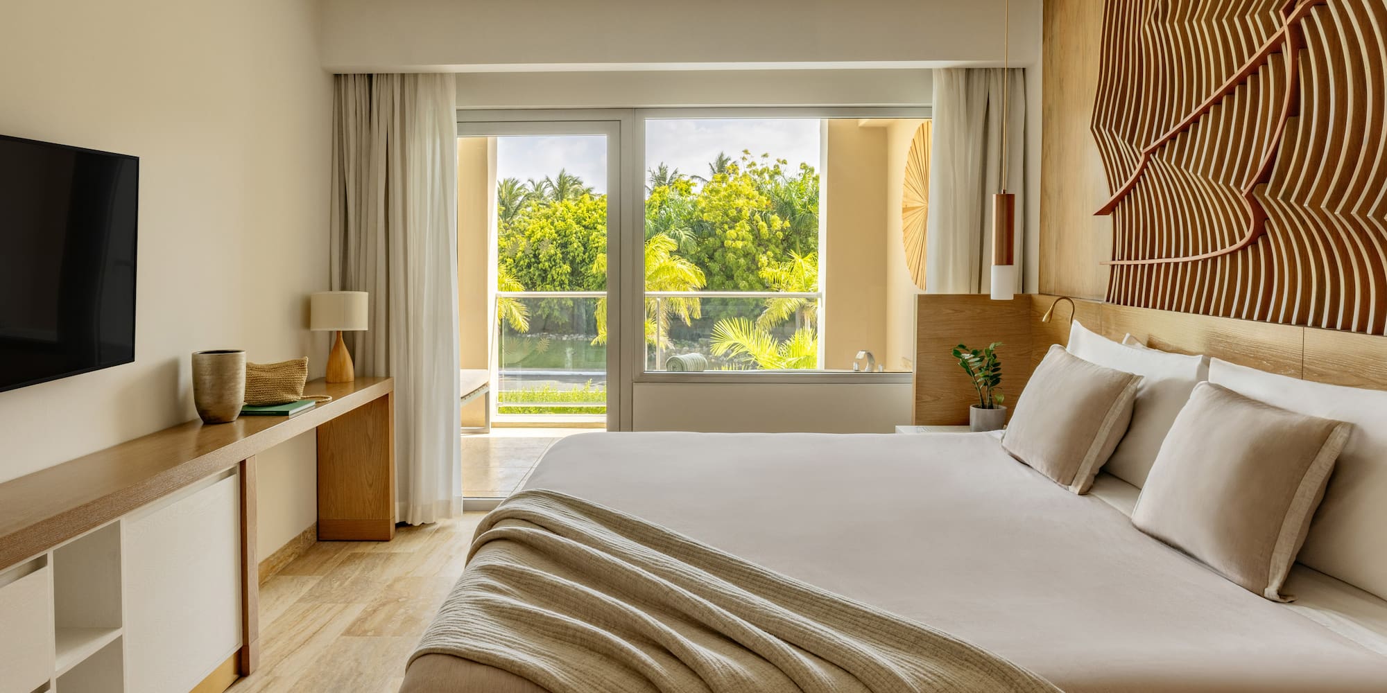 Modern hotel bedroom with king bed, lush tropical view, and decorative wood wall.