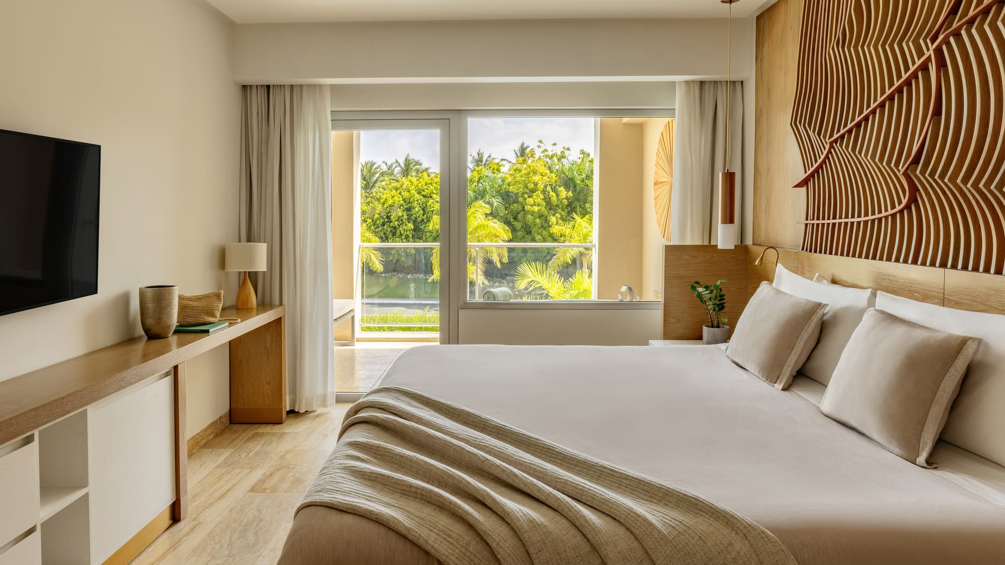 Modern hotel bedroom with king bed, lush tropical view, and decorative wood wall.