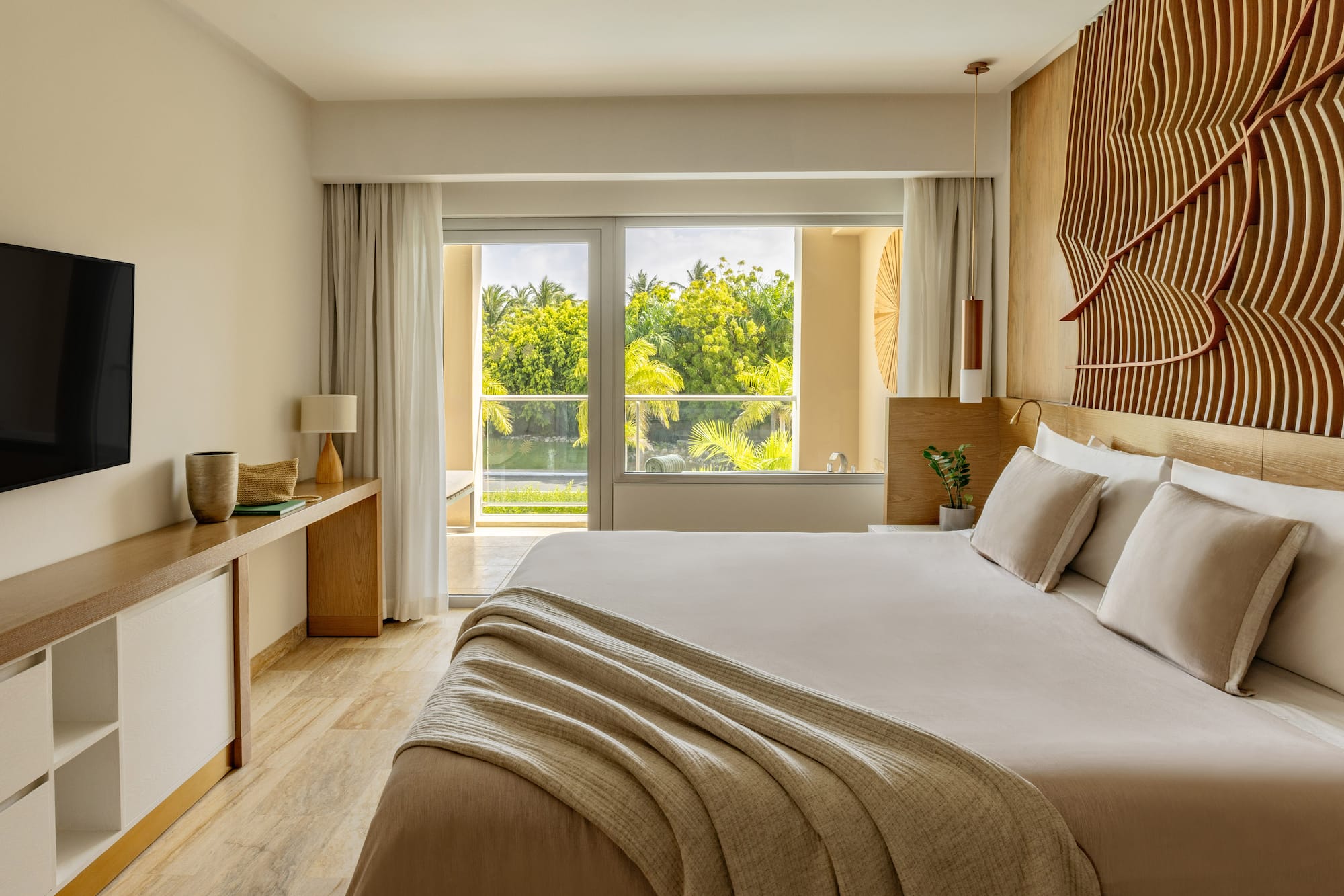 Modern hotel bedroom with king bed, lush tropical view, and decorative wood wall.