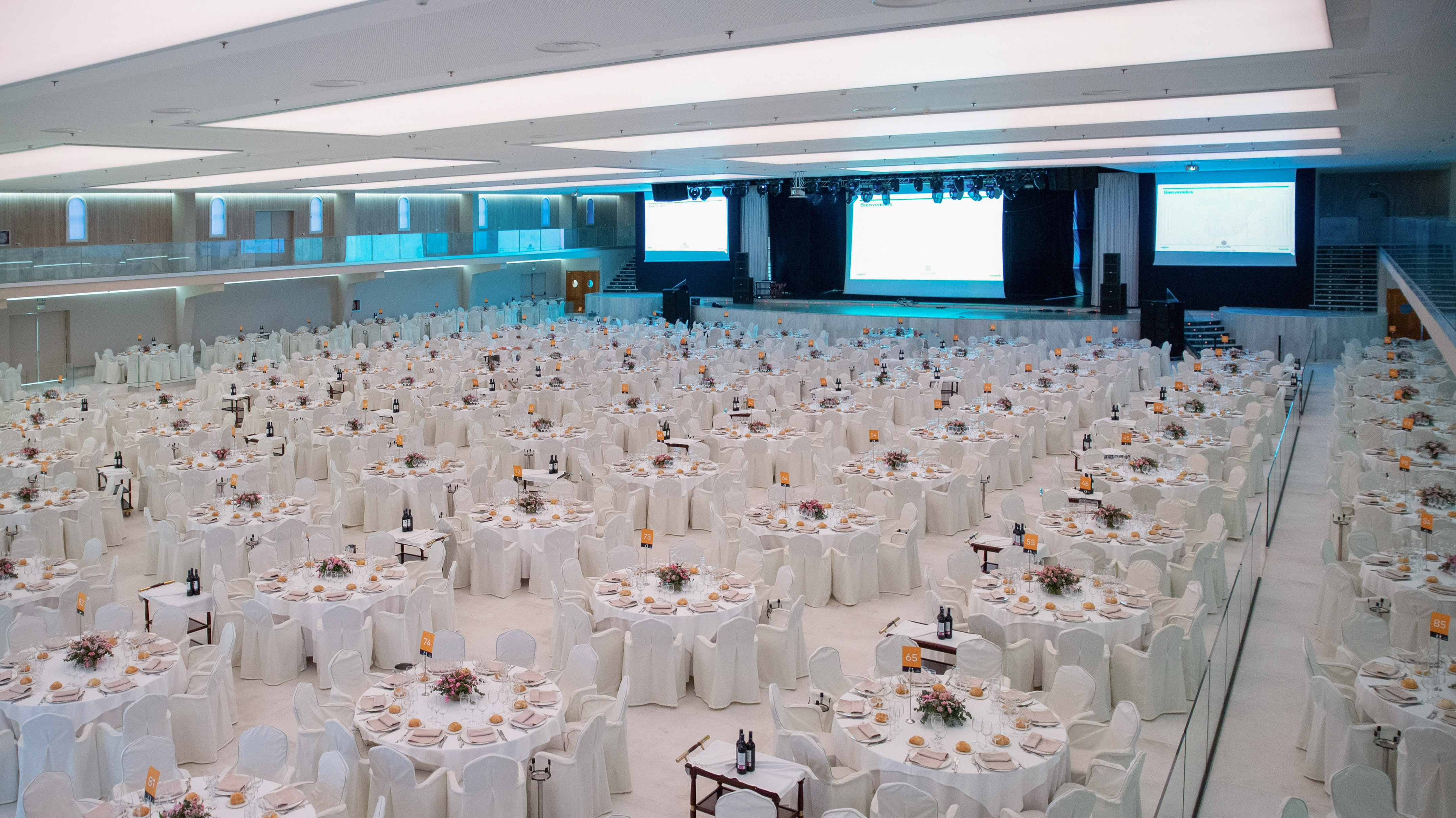 Large banquet hall with set round tables, white chairs, stage, and projection screens.