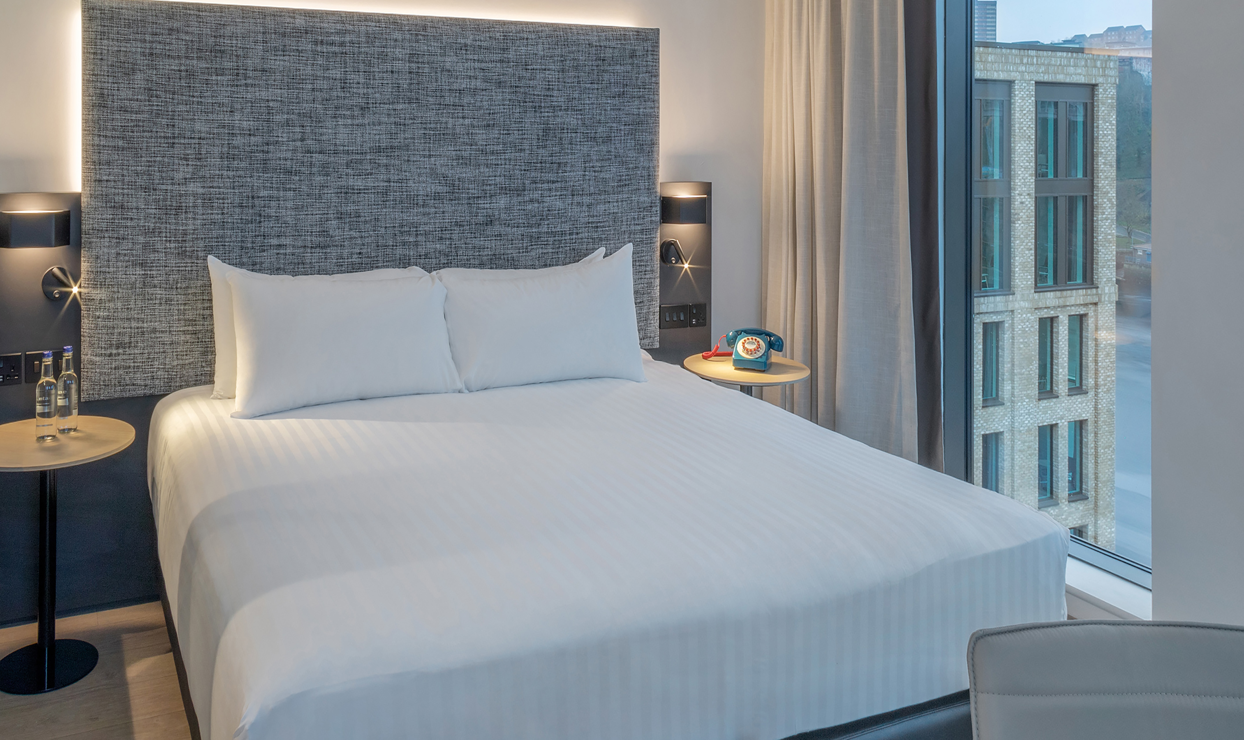 Modern hotel room with white bed, grey headboard, and city view.