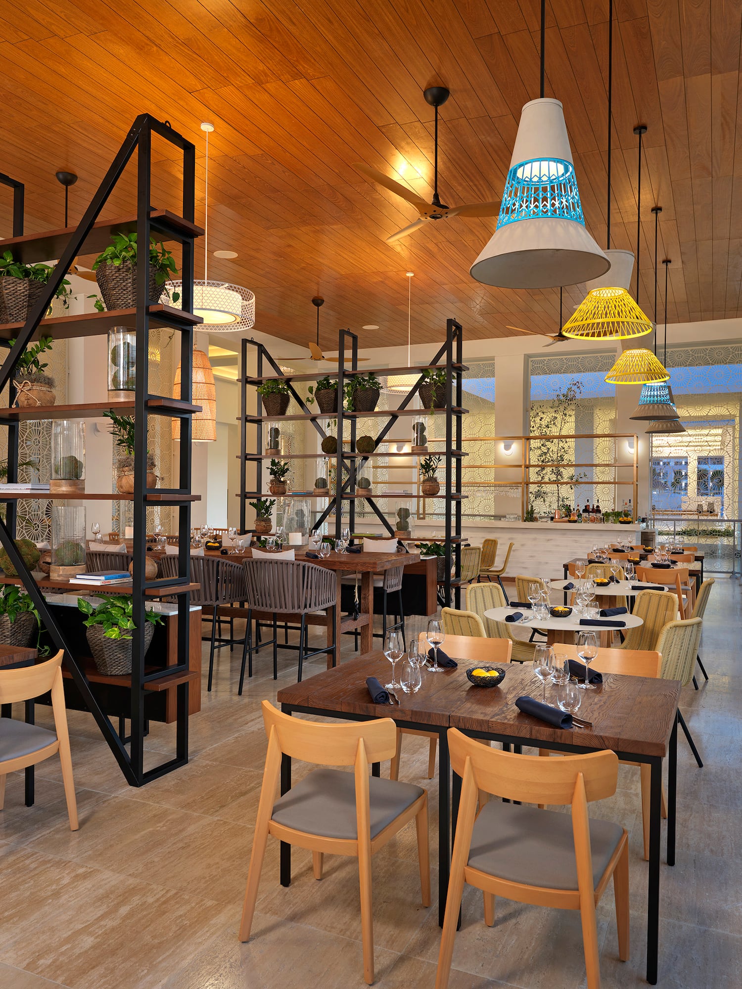 Modern tropical restaurant interior with wood ceiling, plants, and colorful lamps.