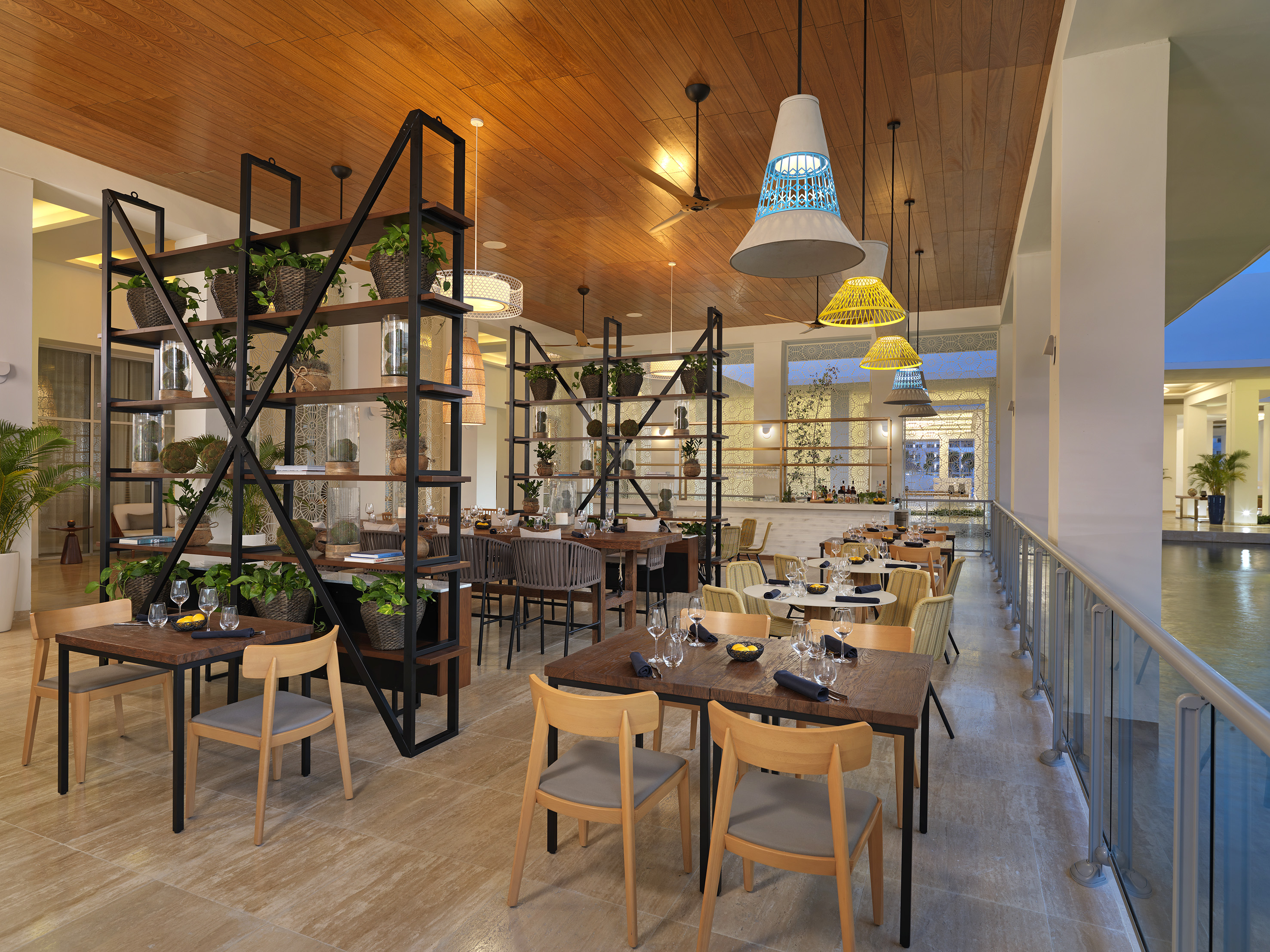 Modern tropical restaurant interior with wood ceiling, plants, and colorful lamps.