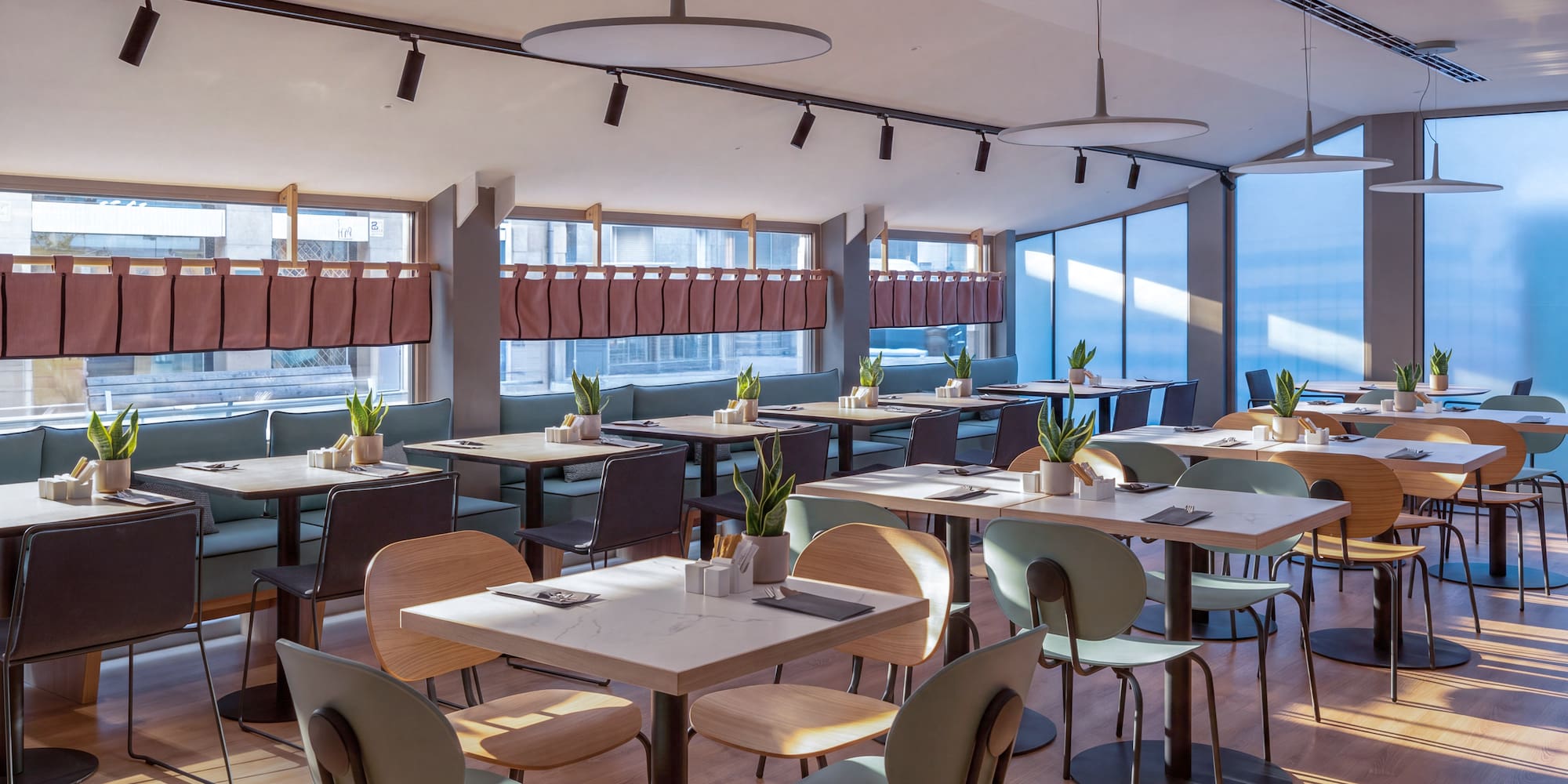 Cafetería moderna y luminosa con mesas, sillas variadas y plantas.