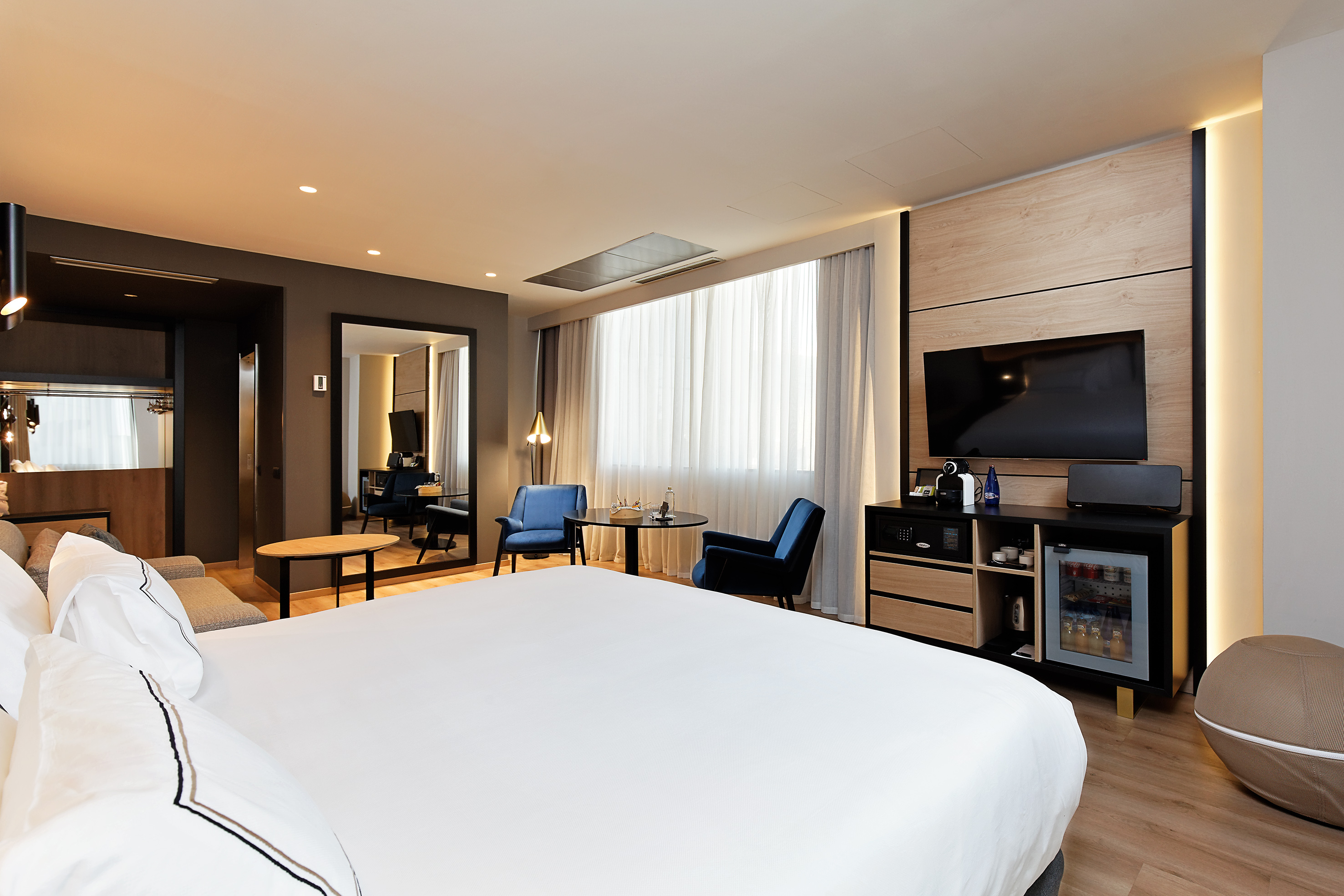 Contemporary hotel room with king bed, flat-screen TV, minibar, and blue armchairs.