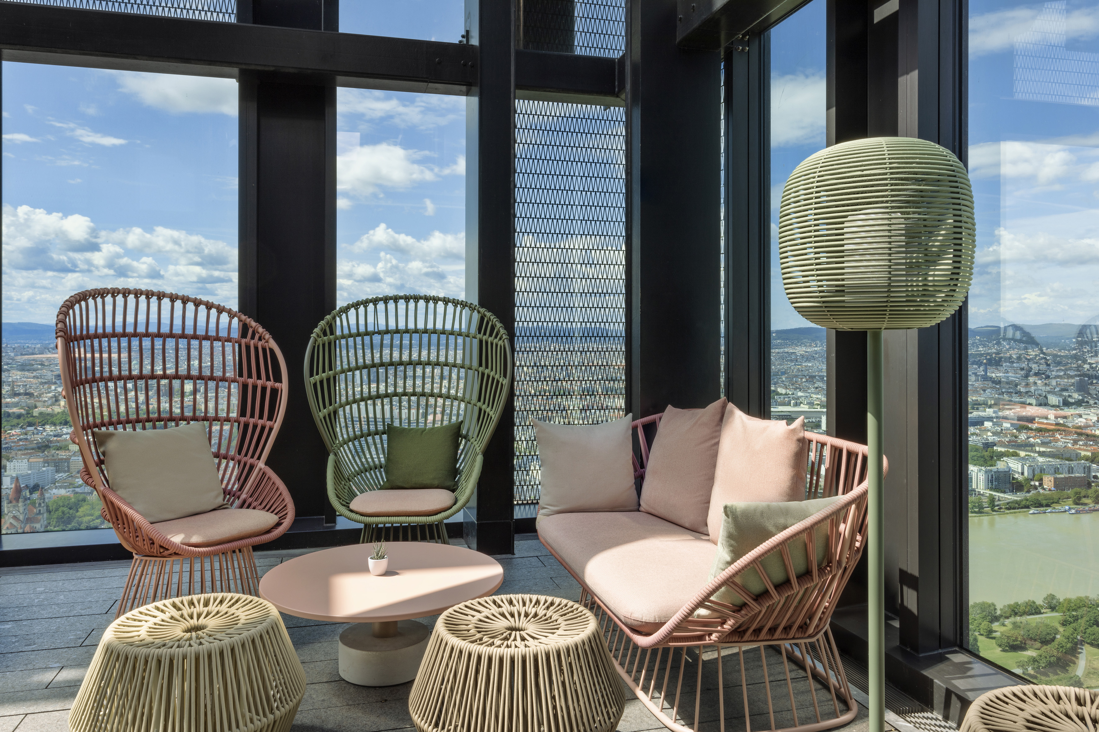 Modern rooftop lounge with pink and green woven patio furniture, city view.
