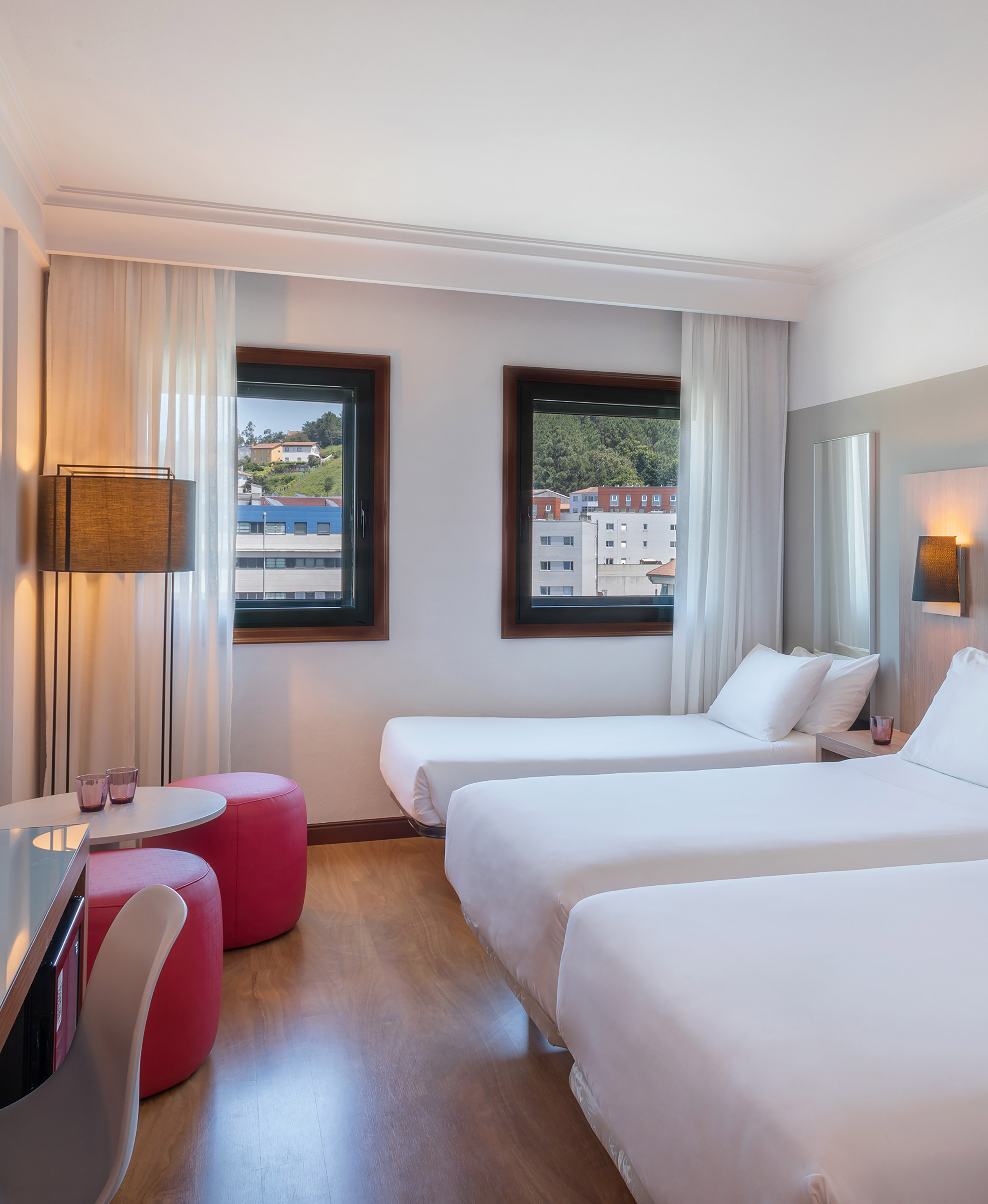 Bright triple hotel room with three single beds, city view, and wood floor.