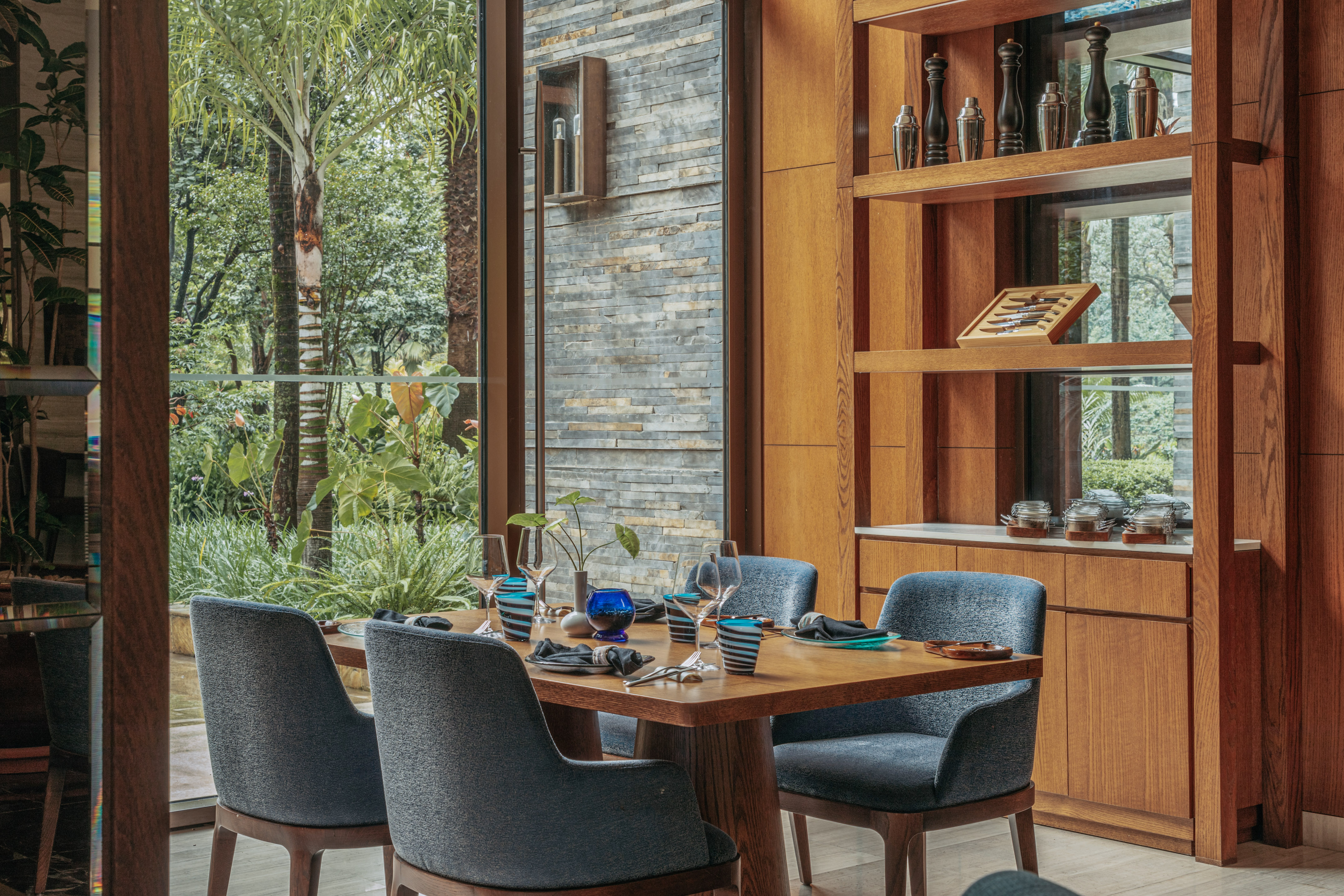 Modern dining room with wooden table, blue chairs, and lush tropical garden view.