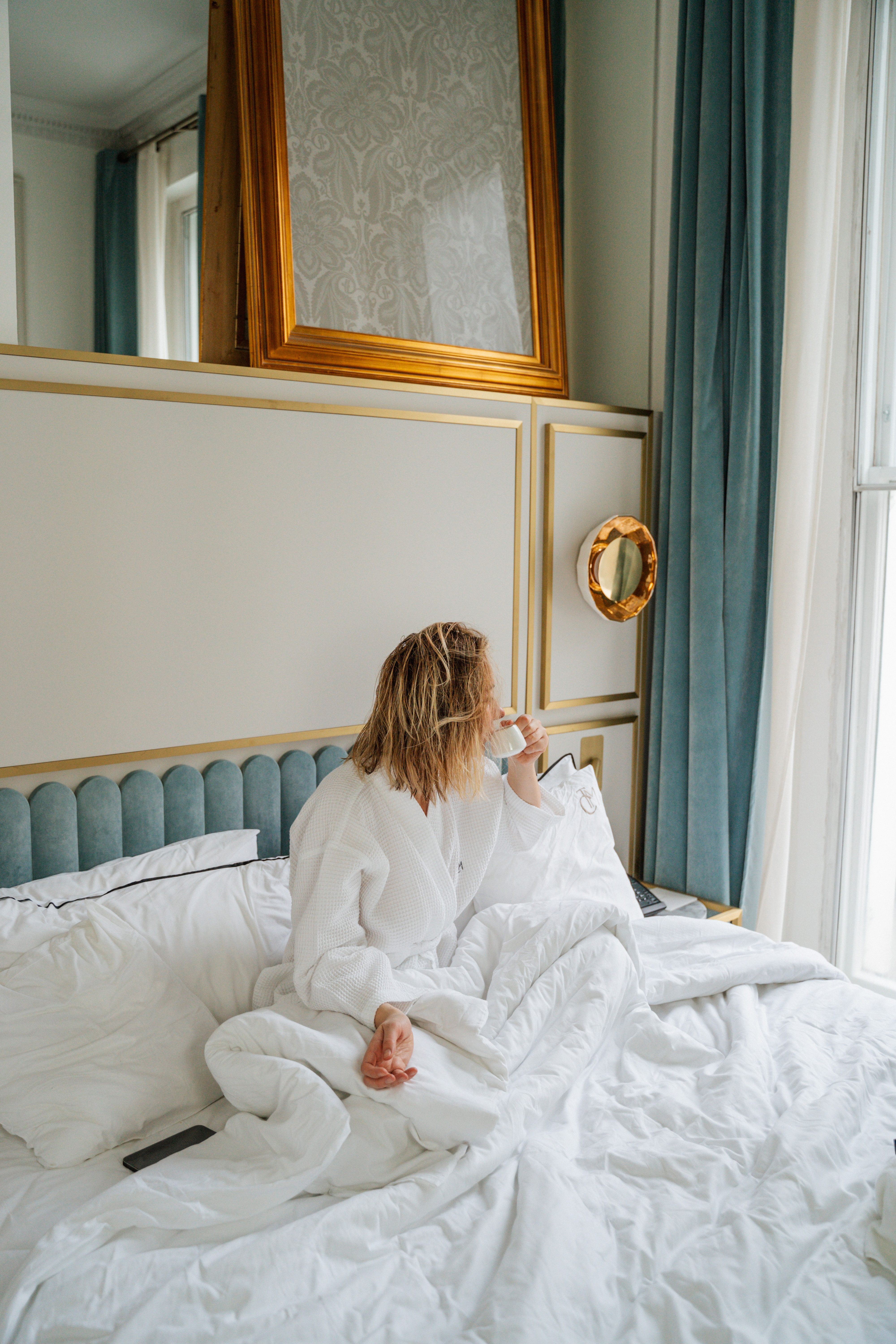 Woman in white bathrobe sipping coffee on a luxury hotel bed.