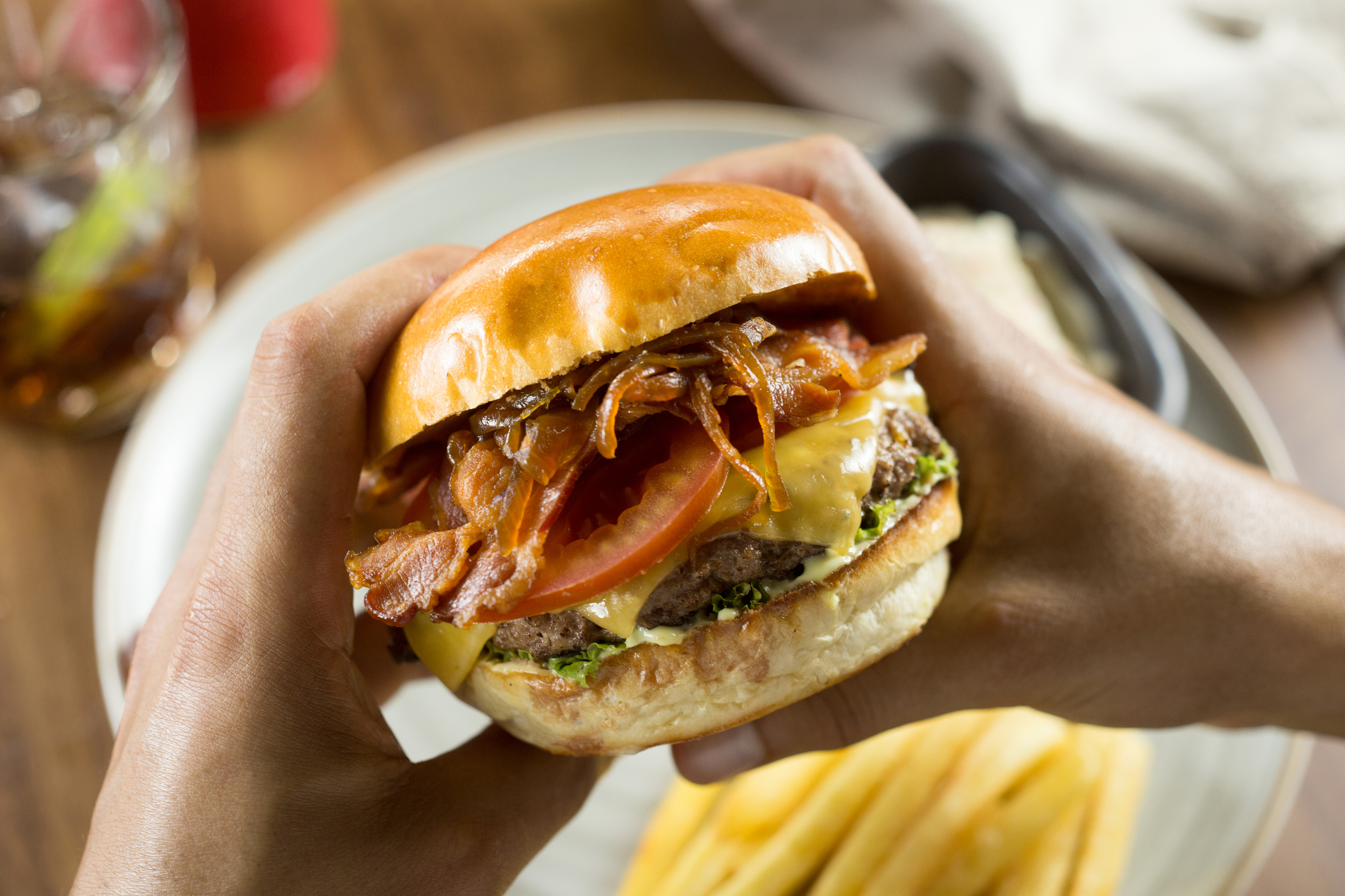 Hands holding a bacon cheeseburger with caramelized onions and fries.