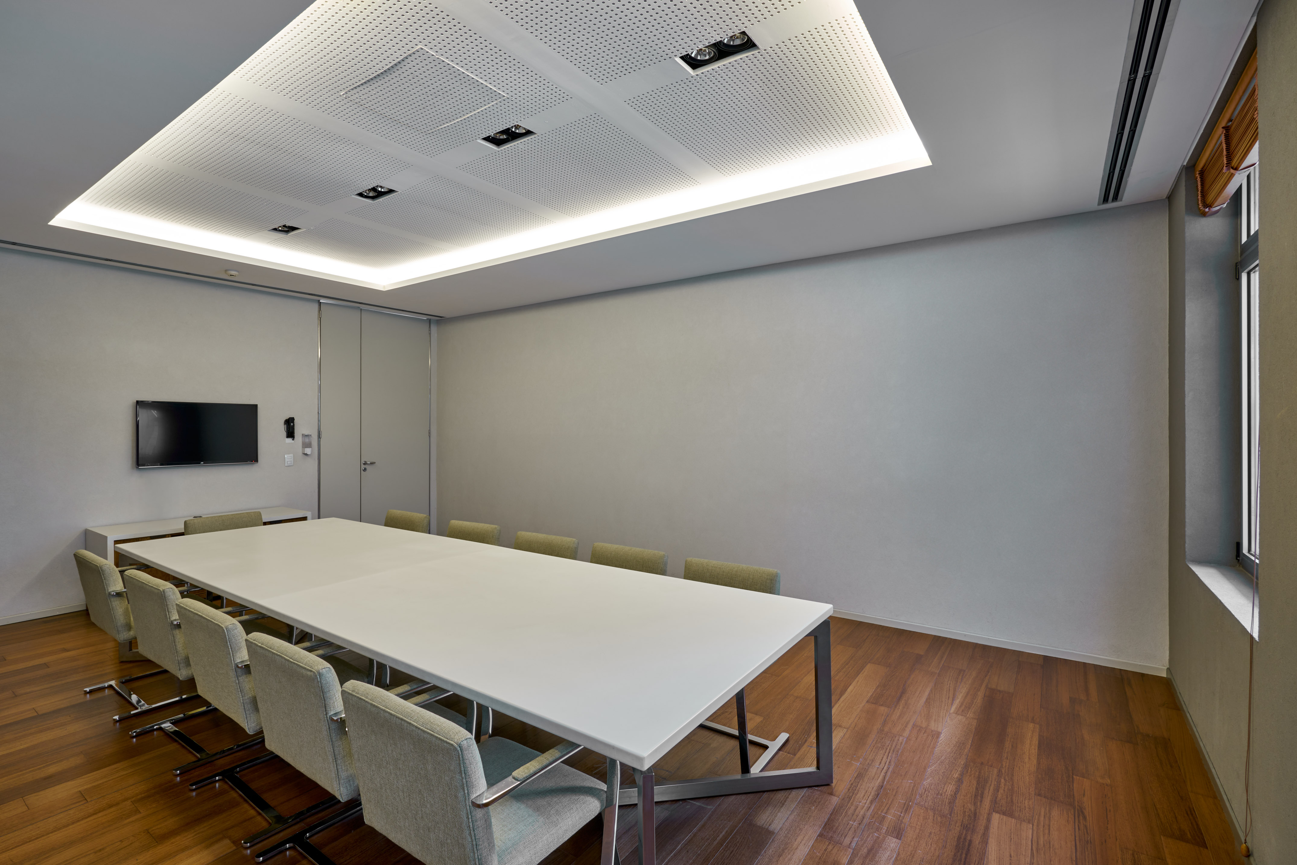 Modern conference room with long white table, grey chairs, wood floor, wall TV.