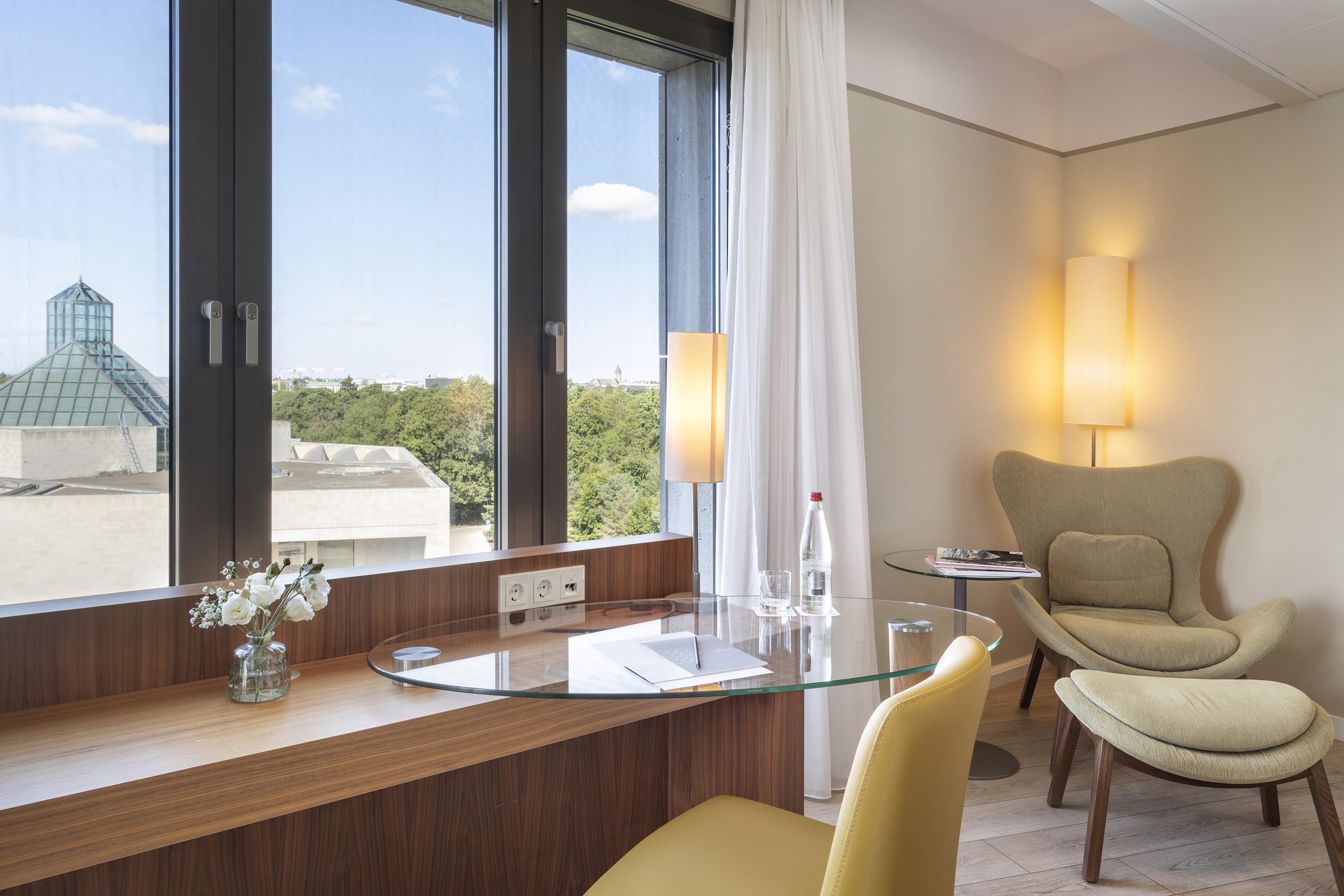 Hotel room with city view, large window, glass desk, and comfy green armchair.