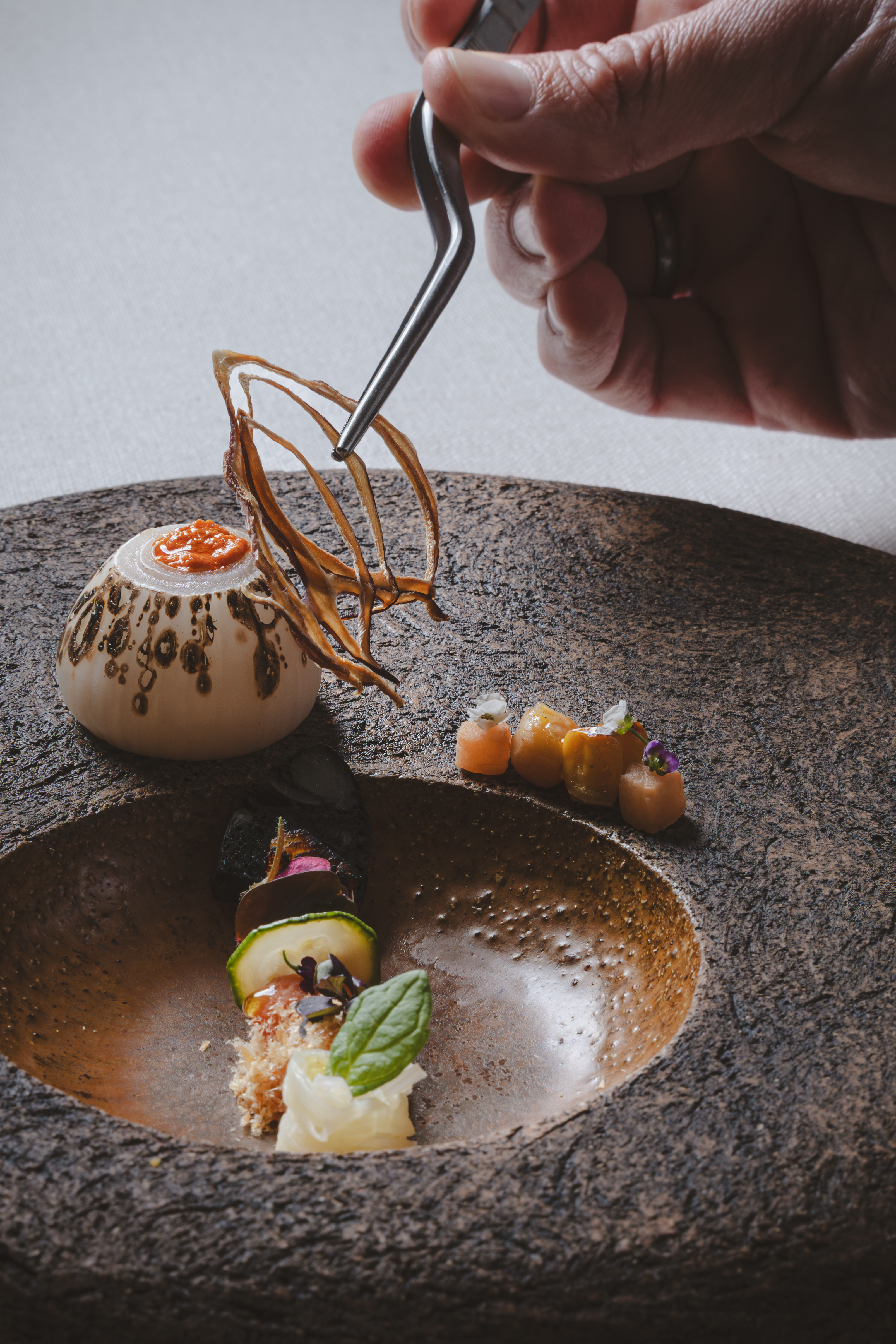 Chef adding crispy garnish with tweezers to gourmet dish on textured plate.