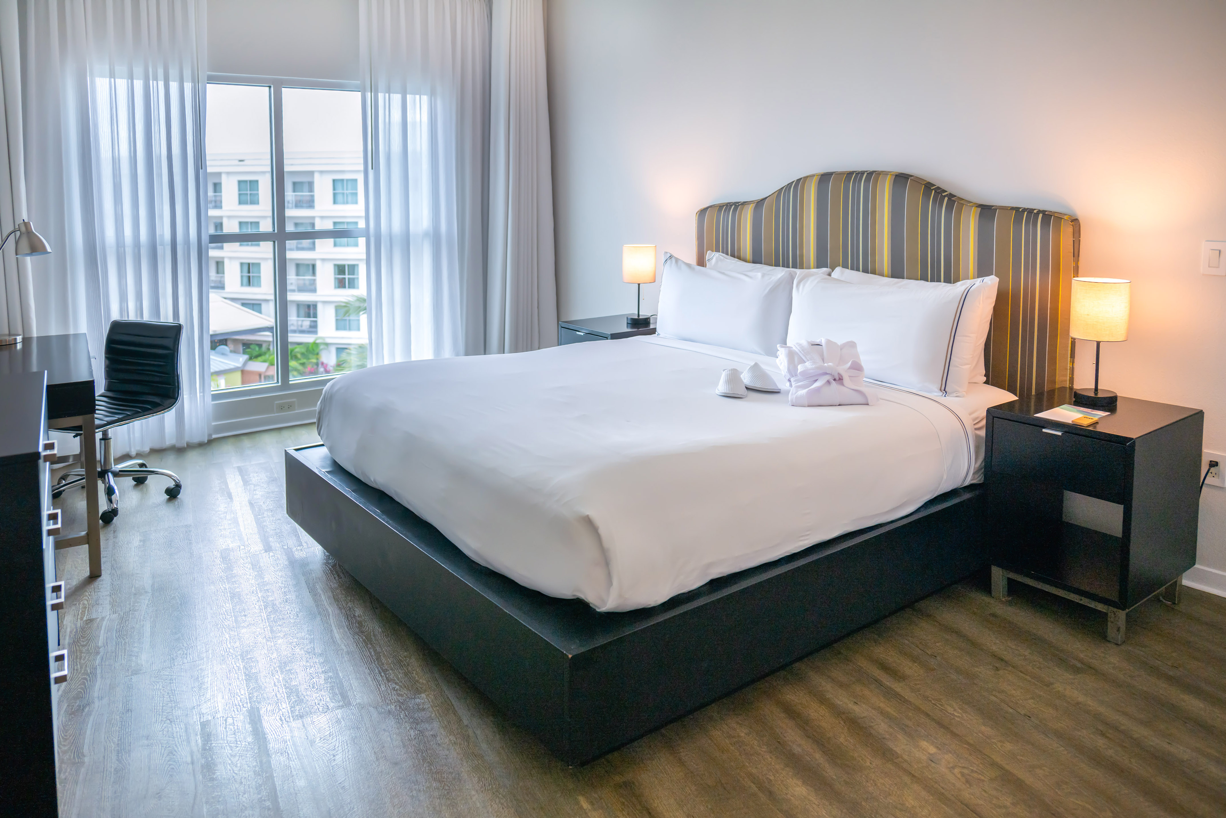 Modern hotel bedroom with large bed, striped headboard, desk, and window view.