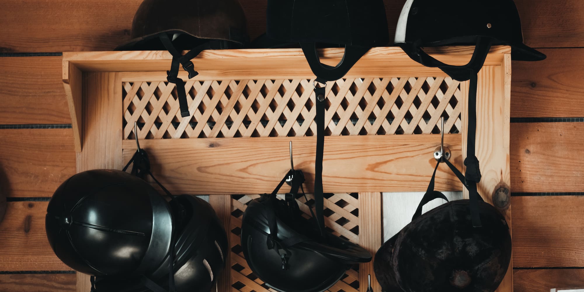 Various horse riding helmets hanging on a wooden shelf in a stable.