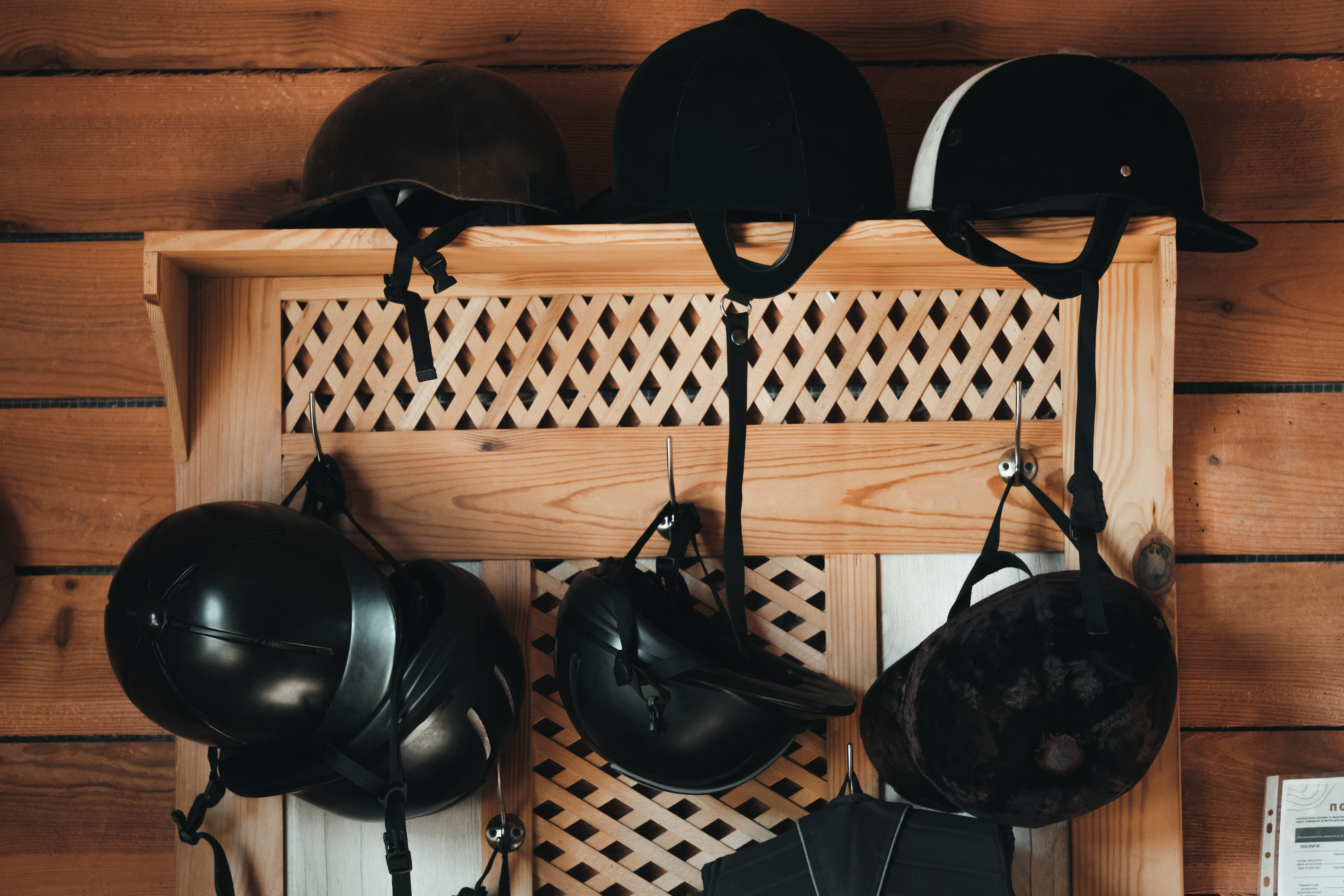 Various horse riding helmets hanging on a wooden shelf in a stable.