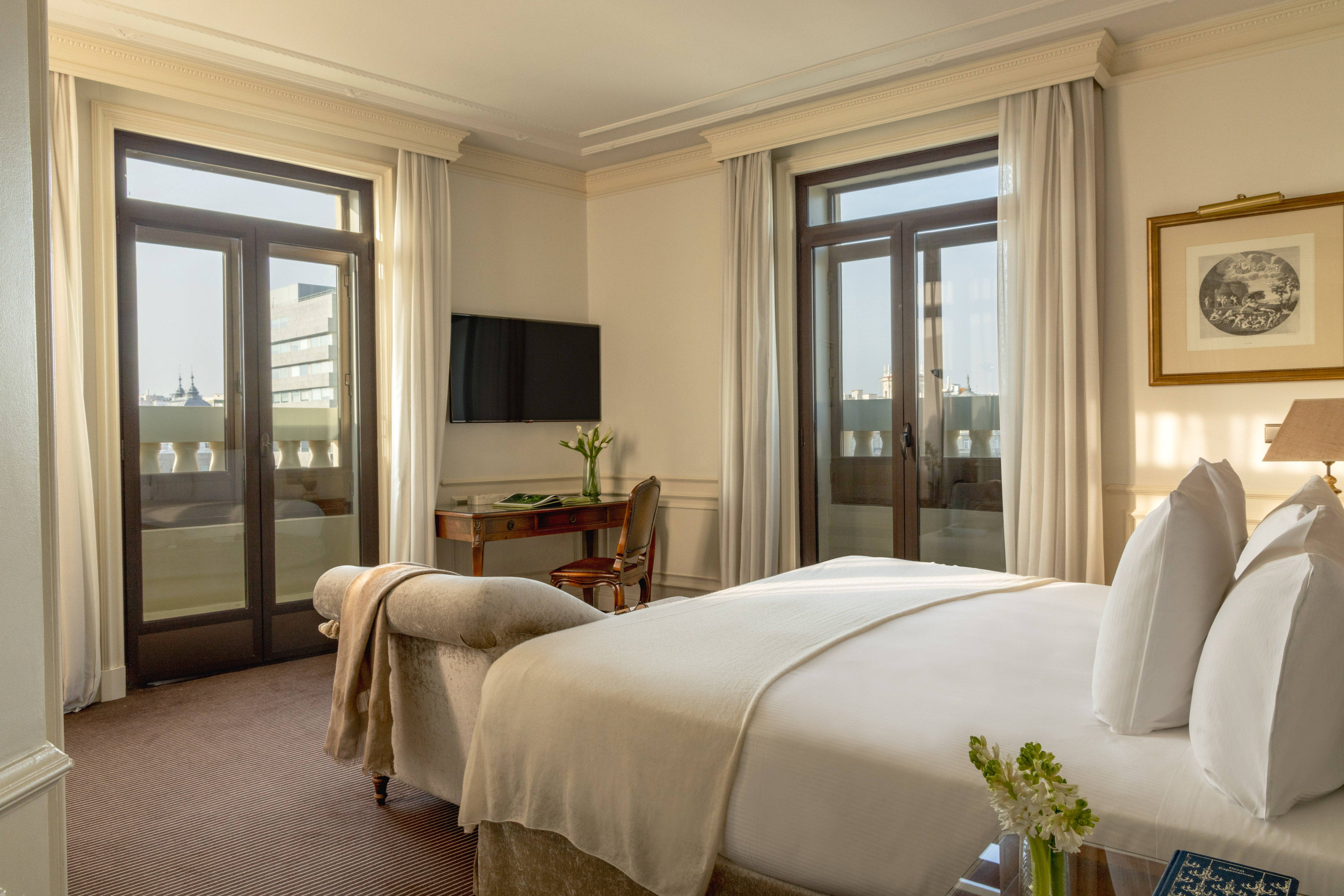 Luxury hotel room with white bed, French doors to balcony, desk, and TV.