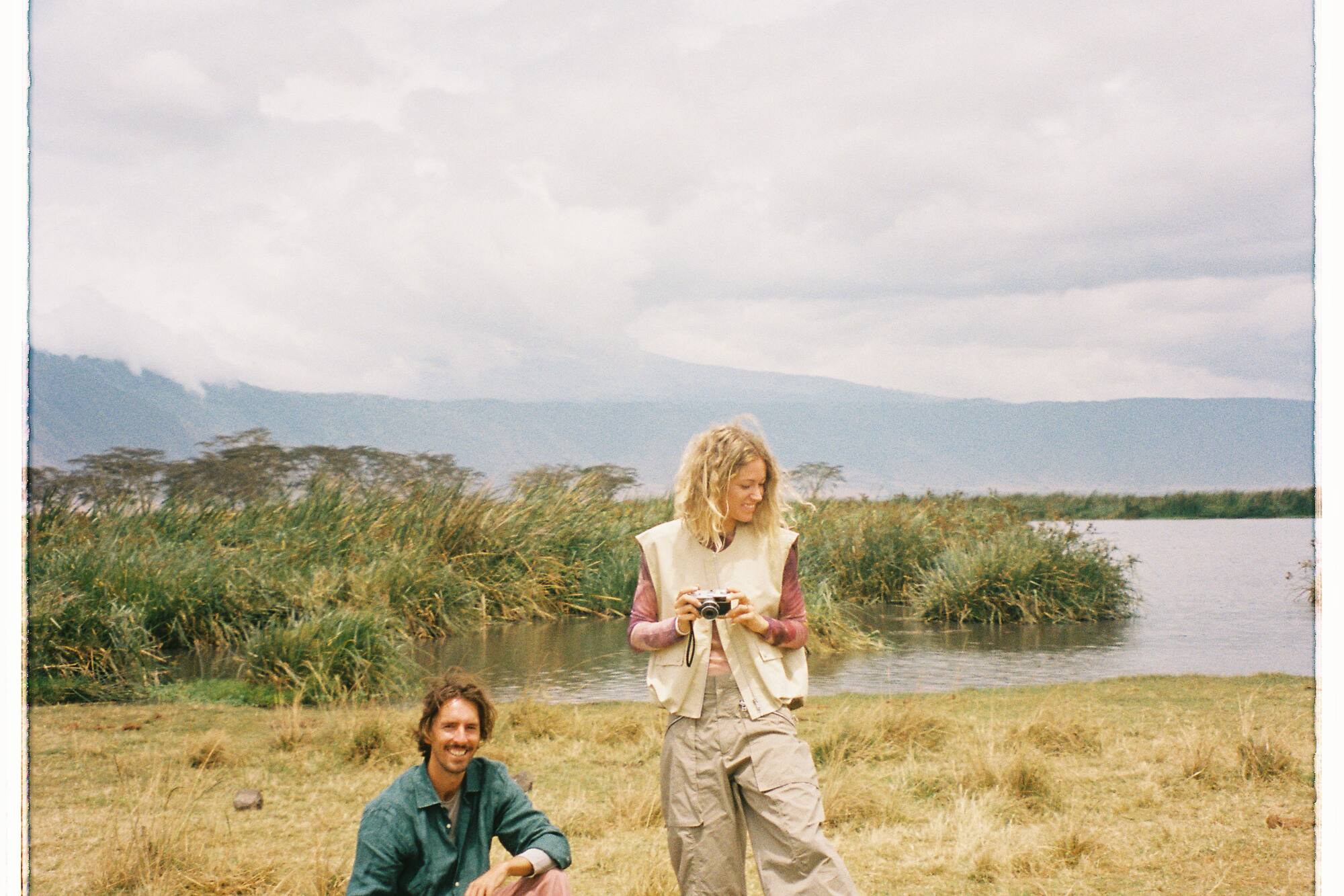 Man sitting, woman with camera by a lake, grassy shore, distant mountains.