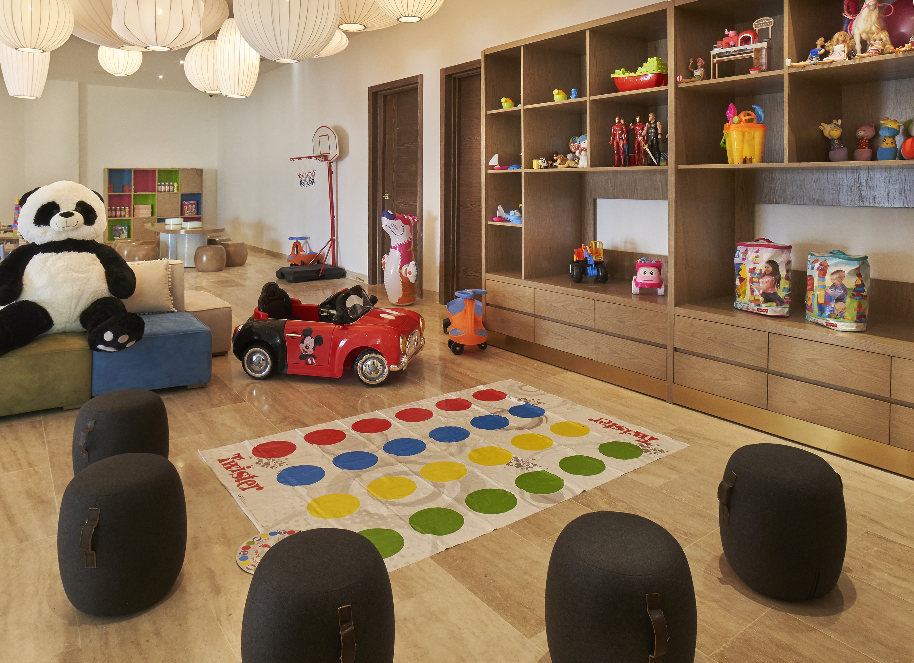 Bright kids' club playroom with panda, ride-on car, Twister mat, and toys on shelves.