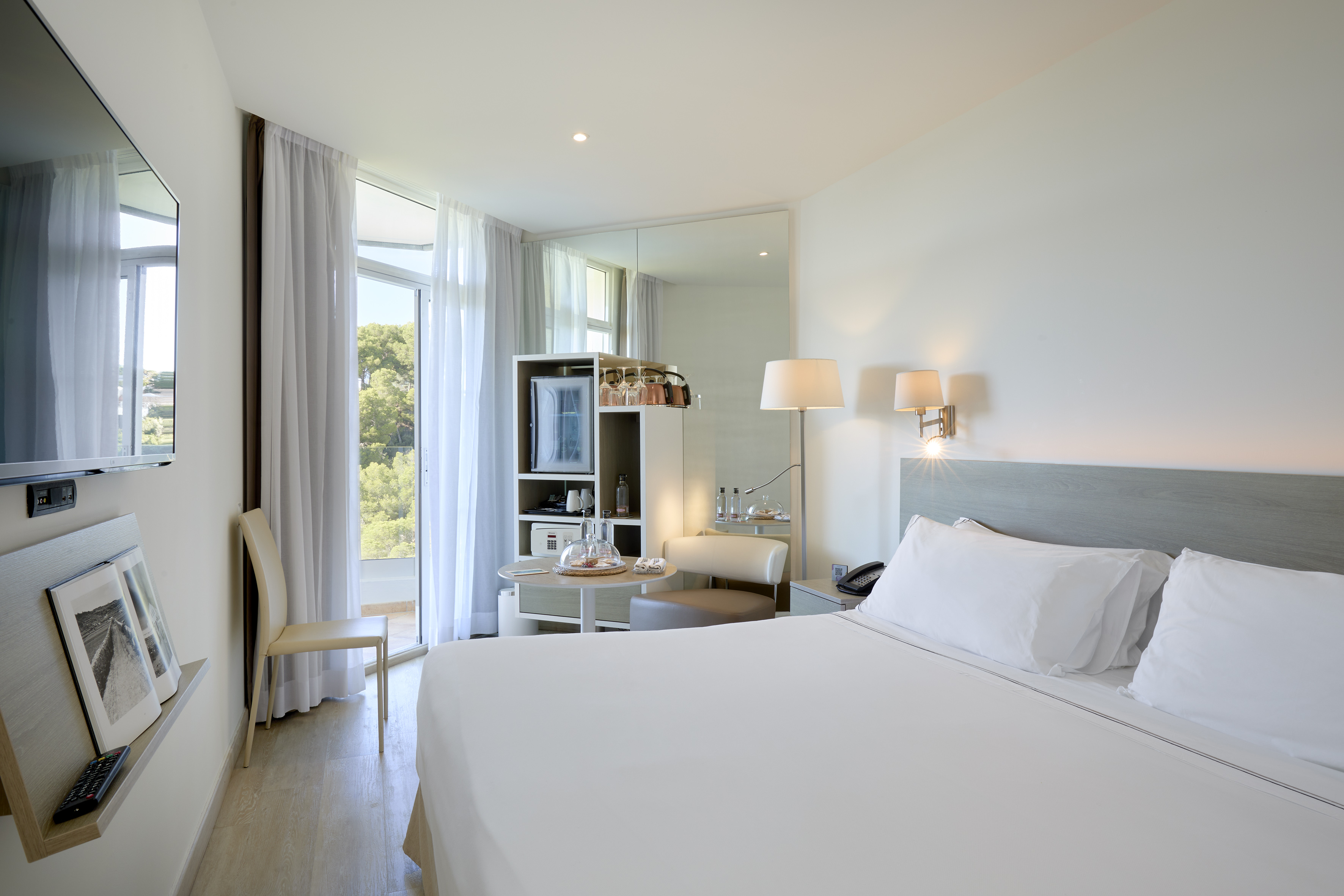 Modern hotel room with white bed, TV, minibar, and a bright balcony view.