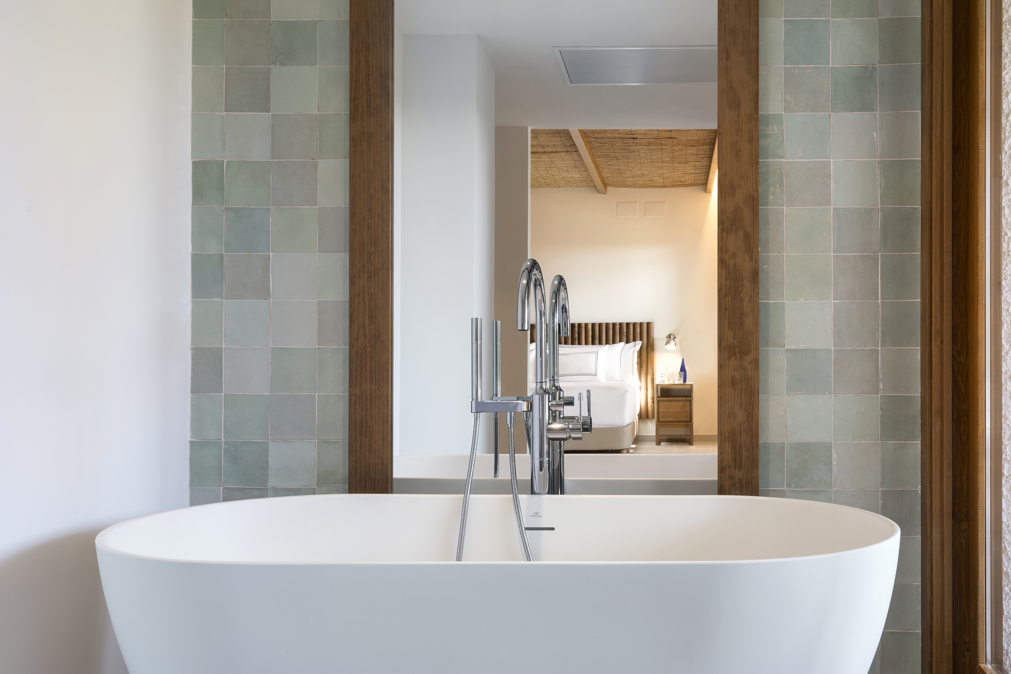 Freestanding bathtub with modern faucet, green tiled wall, and bedroom view.