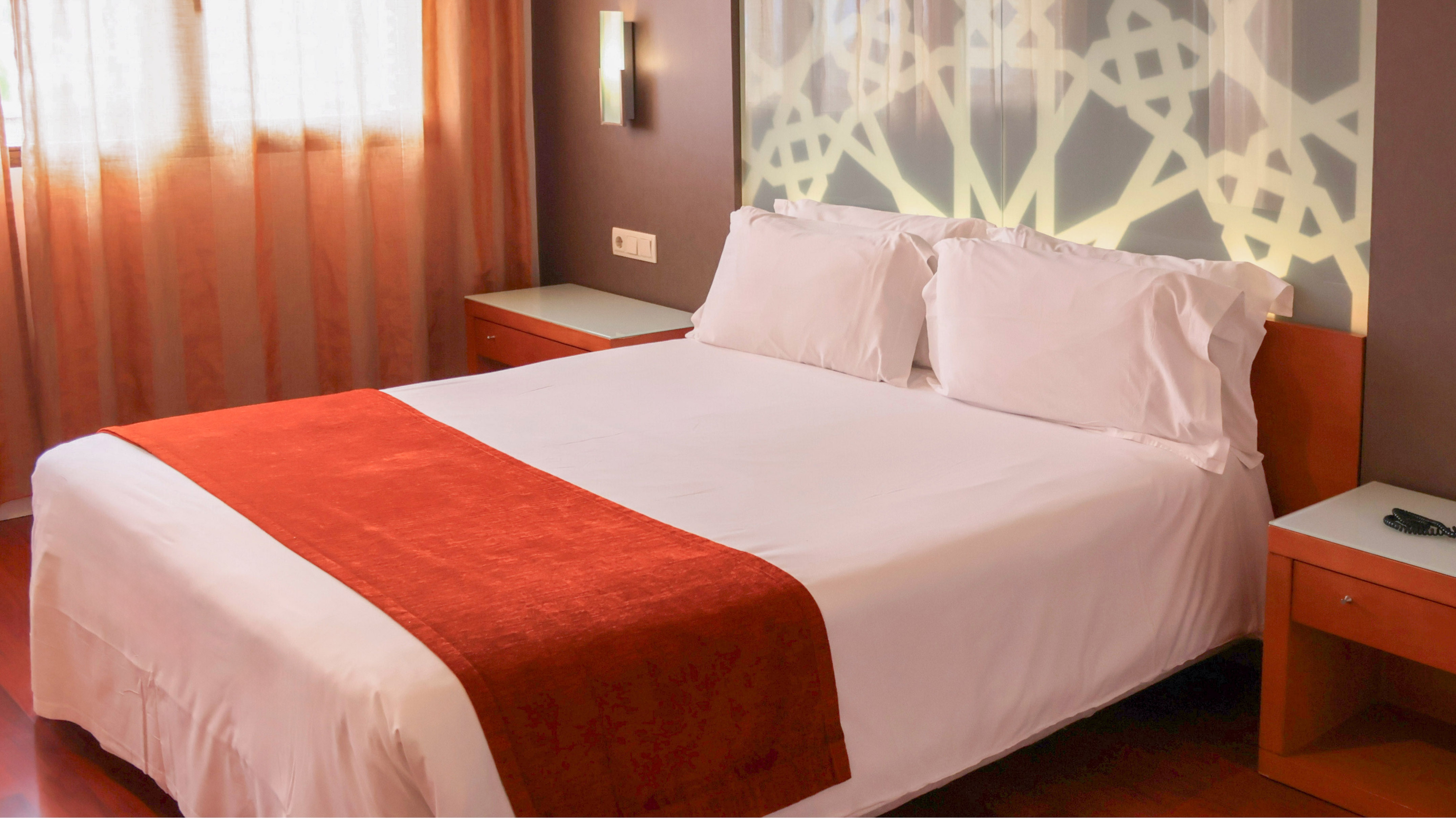 Hotel room bed with white linens, orange runner, and geometric headboard.