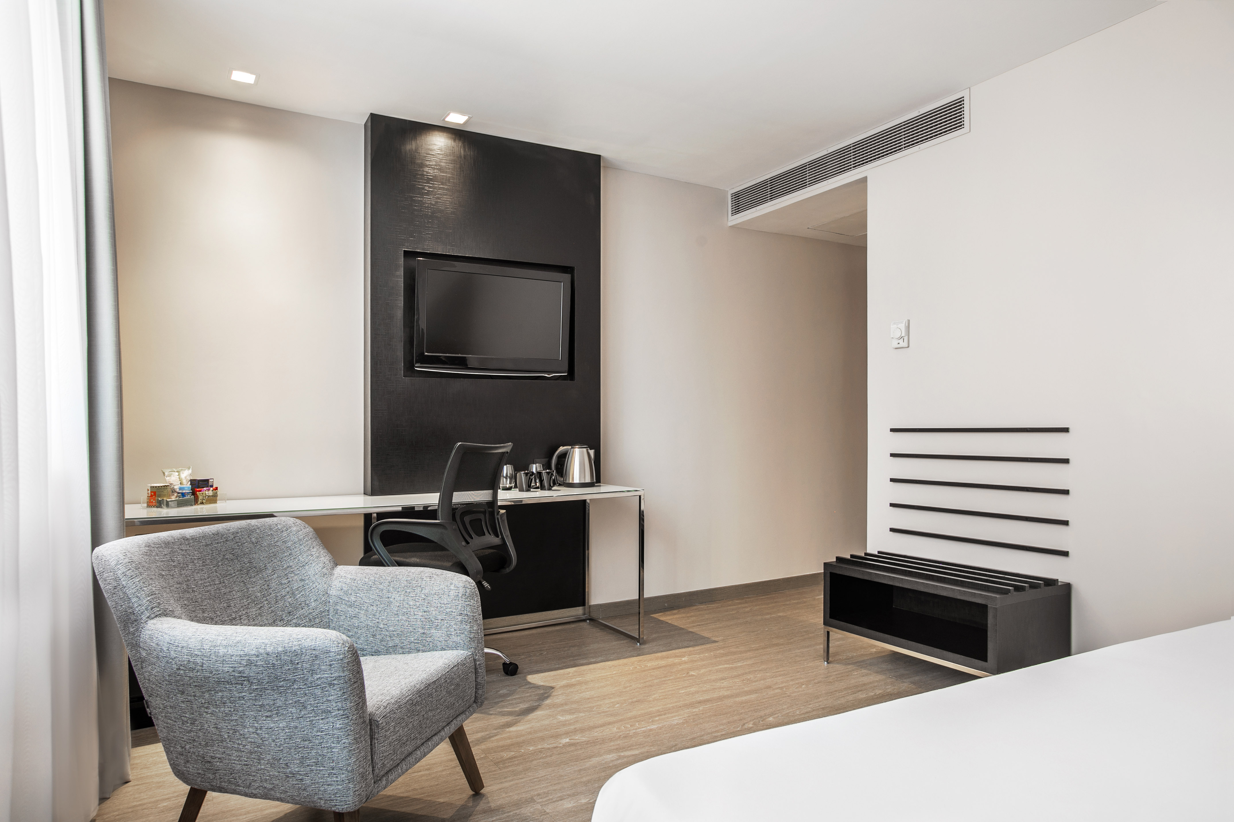 Contemporary hotel room featuring a desk, TV, armchair, and luggage bench.