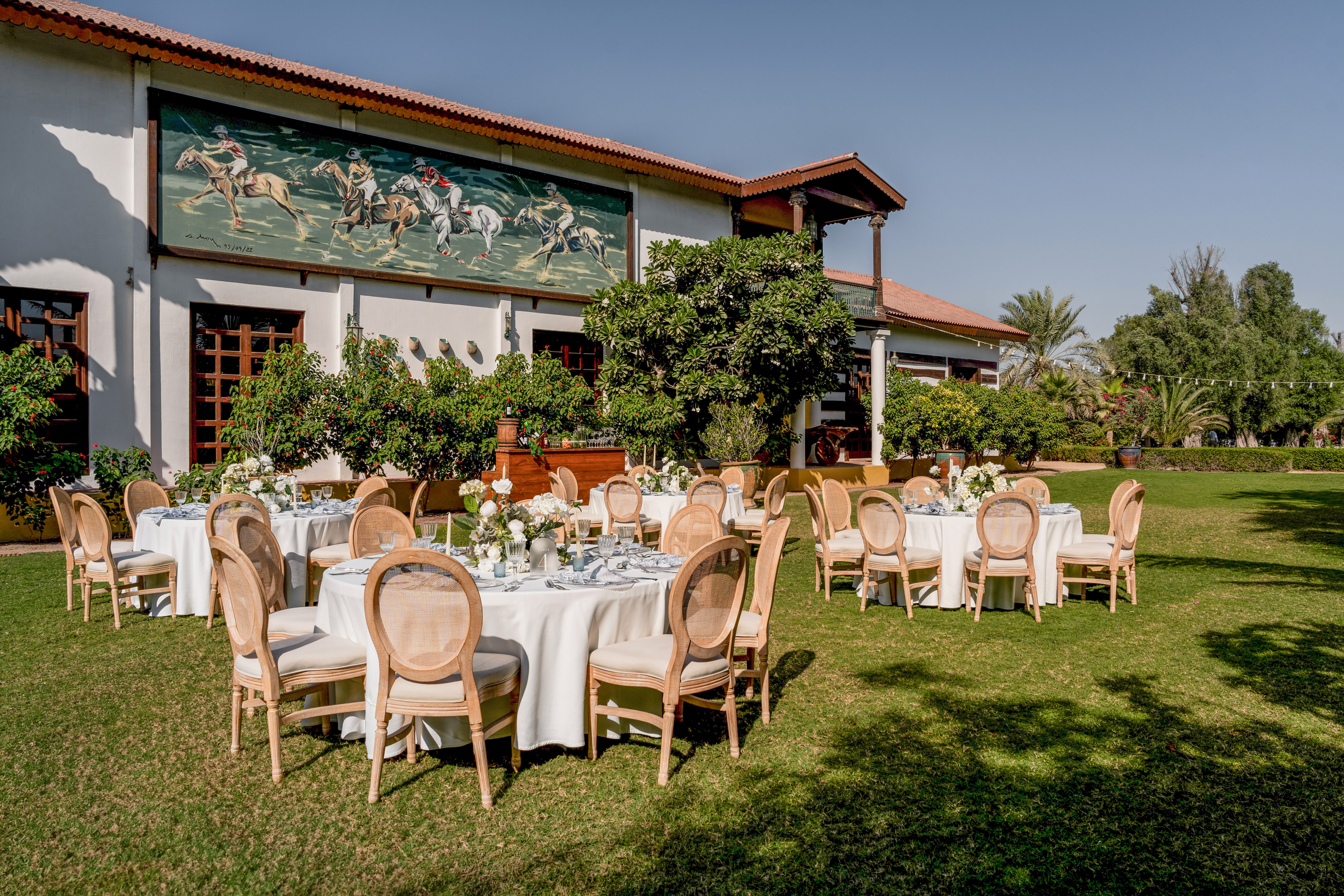 Elegant outdoor event tables on lawn, beside building with large polo mural.