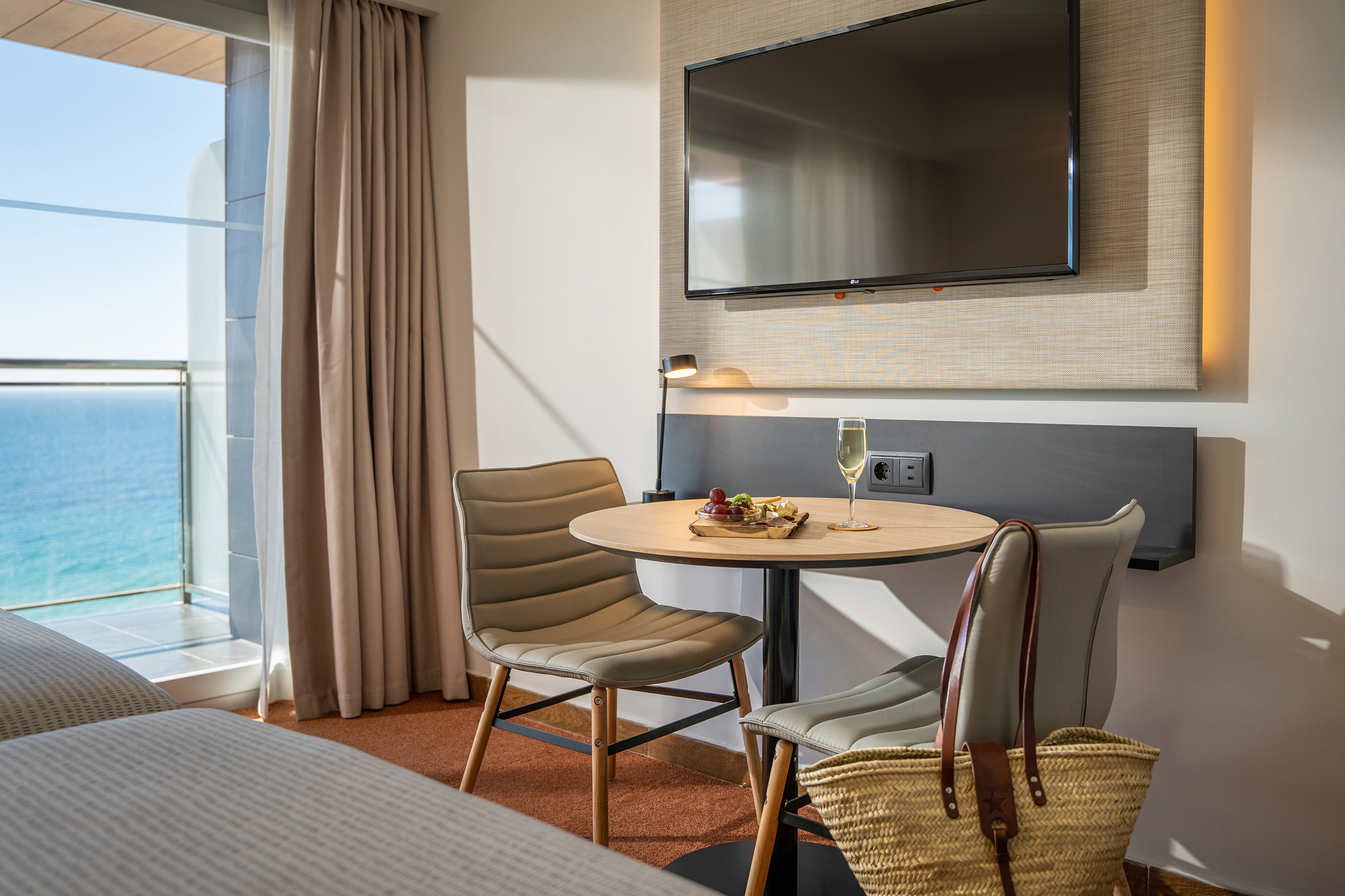 Ocean view hotel room with dining table, champagne, and wall TV.