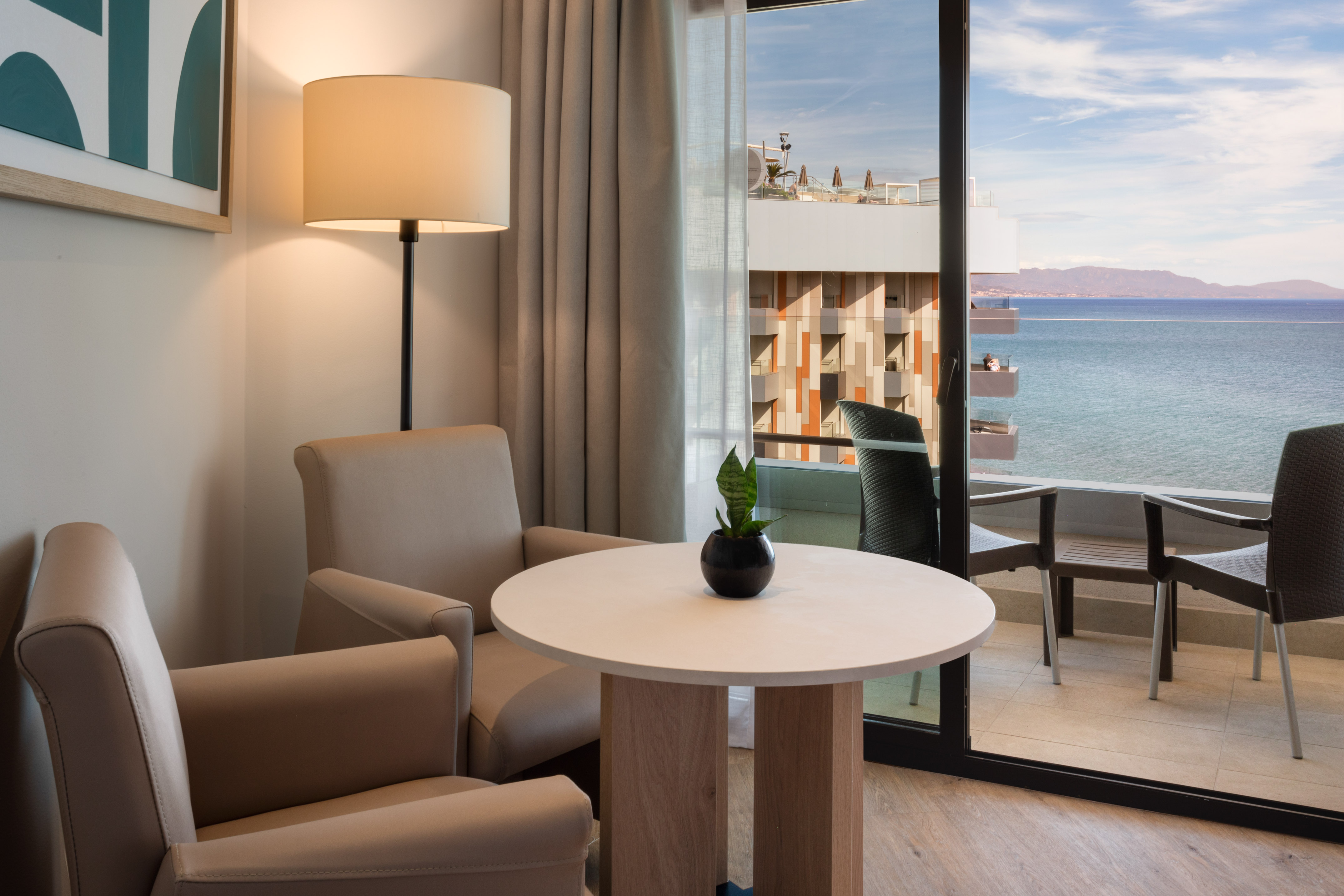 Hotel room with ocean view balcony, armchairs, and a round table.