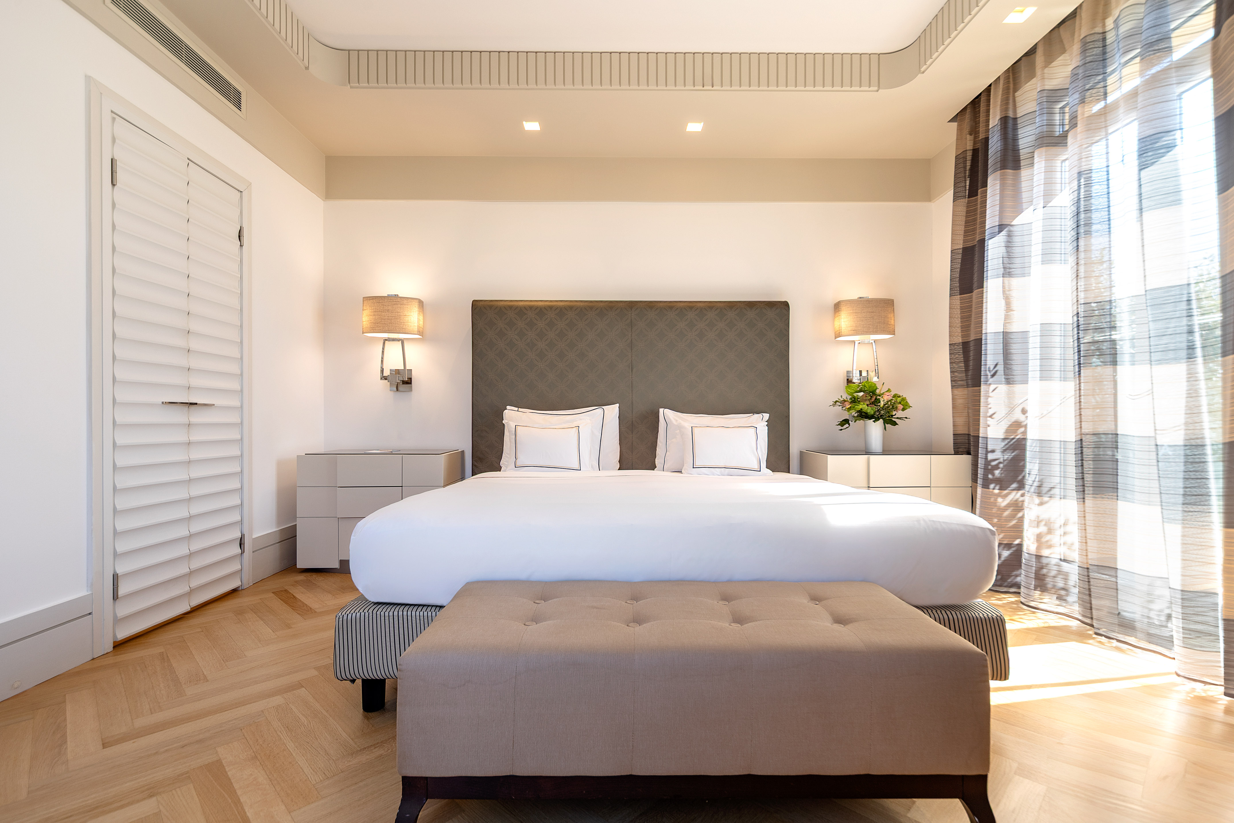 Sunny hotel bedroom with large bed, elegant headboard, and herringbone floor.